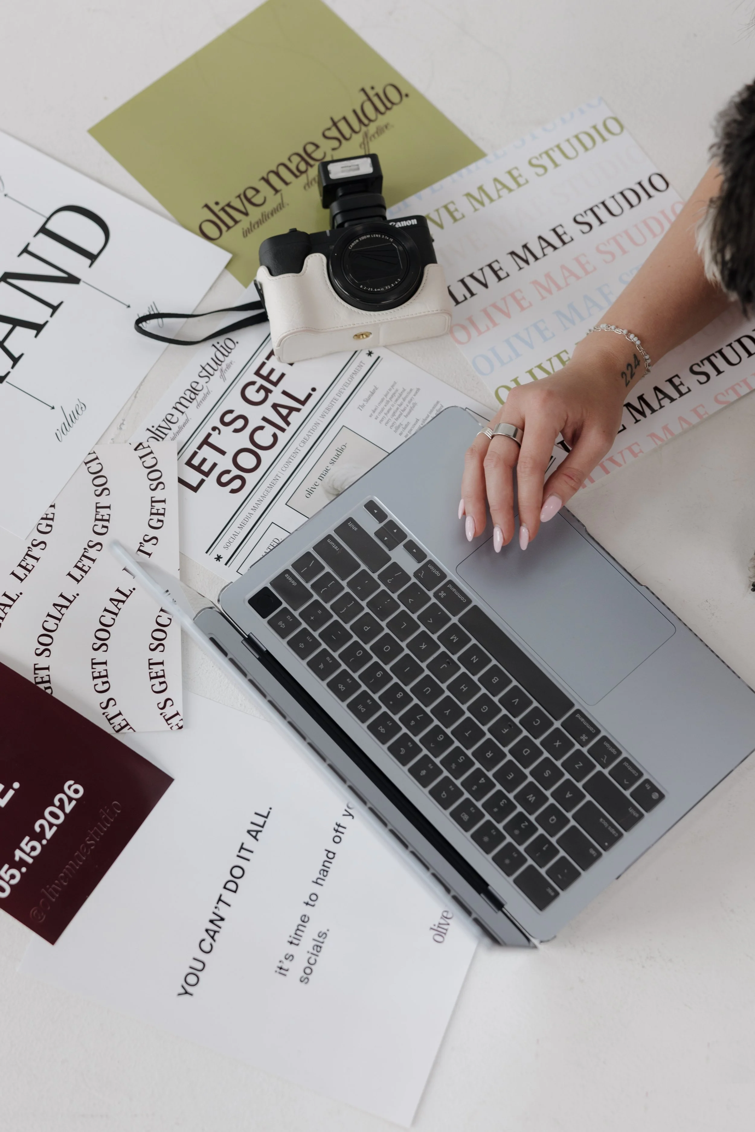 A workspace with promotional materials for Olive Mae Studio, including printed flyers and posters, a camera, a laptop, and a person's hand with rings and a bracelet.