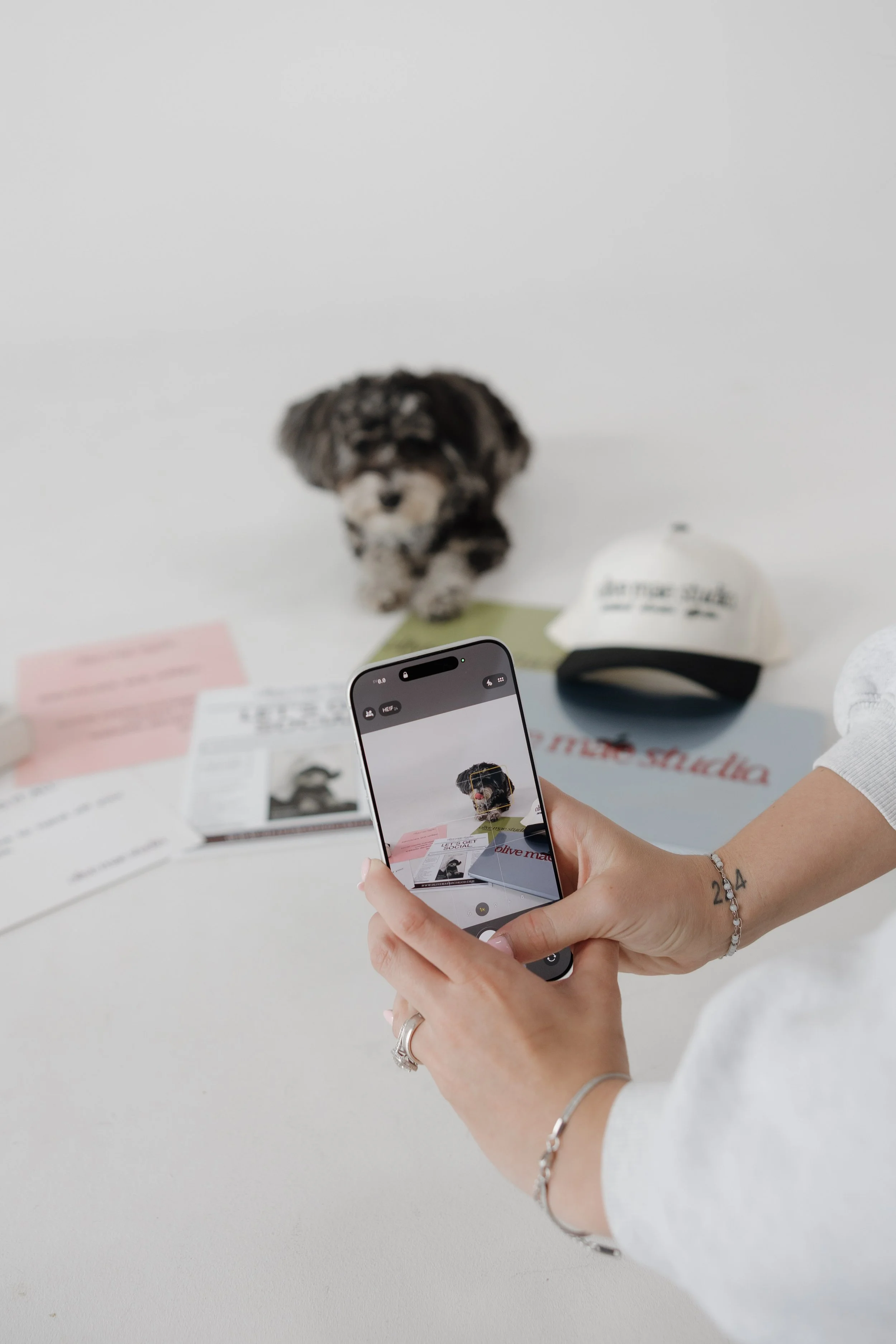 A person taking a photo of a small dog figurine with a smartphone. The figurine is placed on a table surrounded by papers, a cap, and other objects.