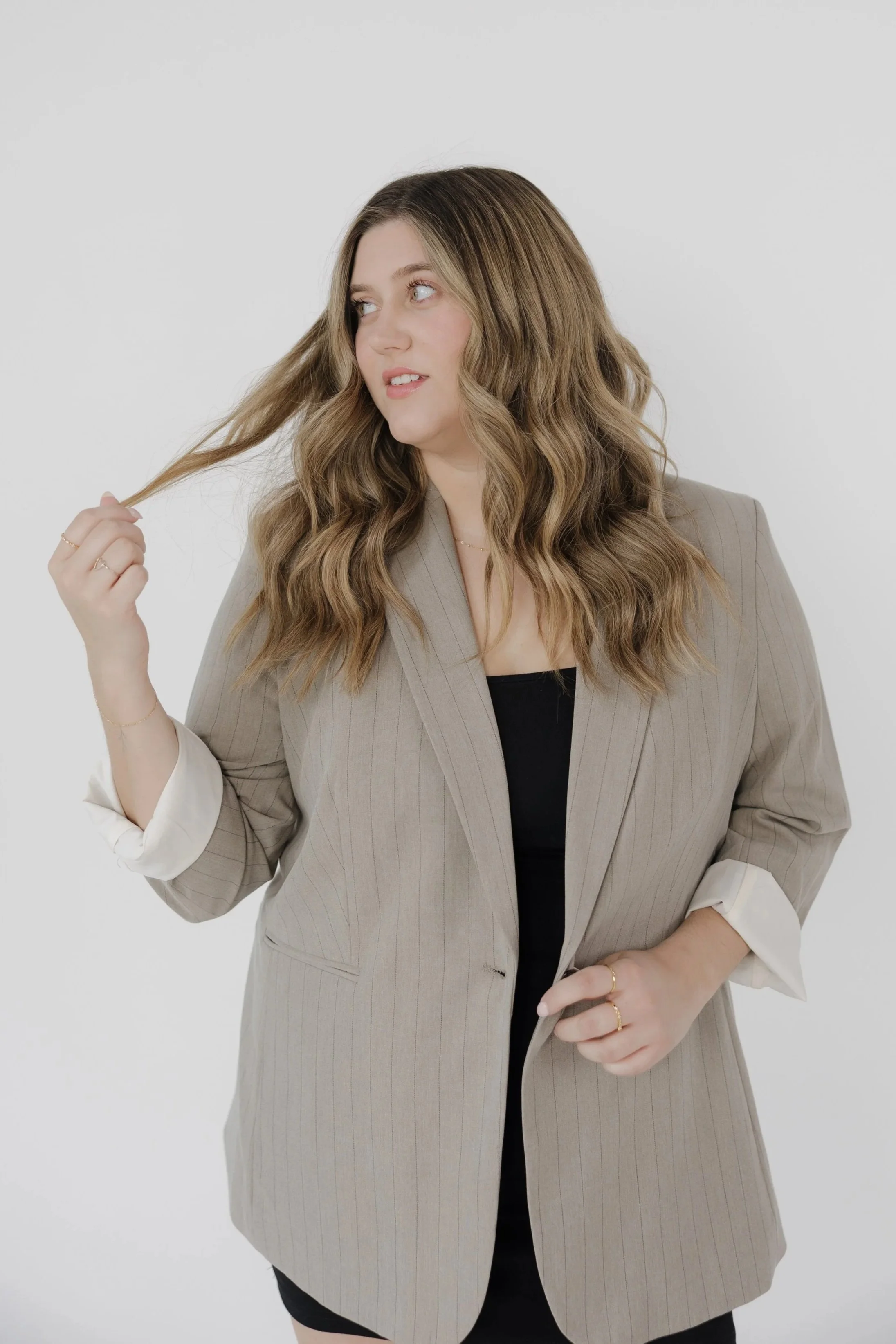 A woman with long, wavy, light brown hair wearing a beige pinstripe blazer over a black top, holding a strand of hair with her right hand and looking to her left, standing against a plain white background.