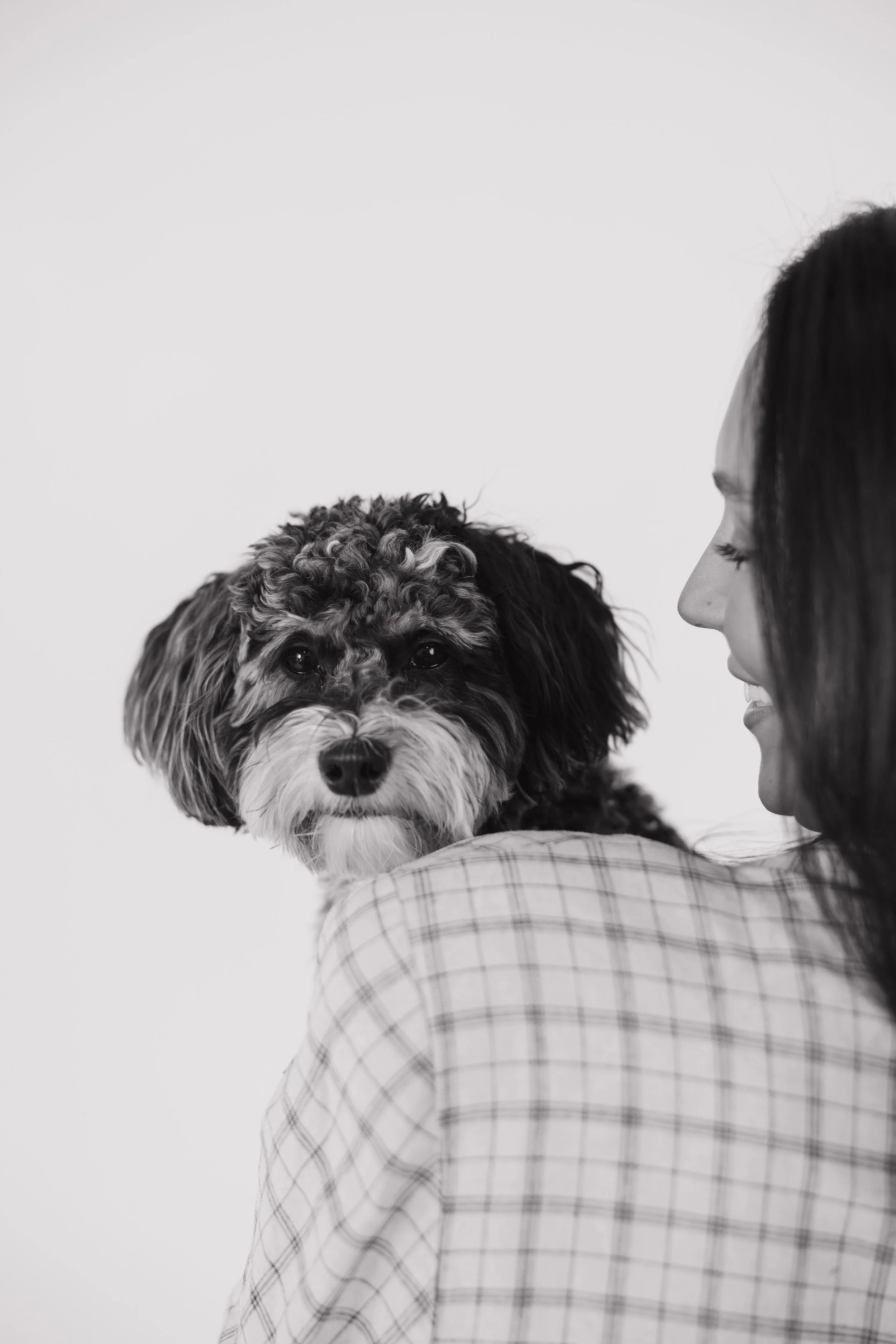 A woman with long hair and a plaid shirt holding a small dog with curly fur looking at the camera, against a plain background.