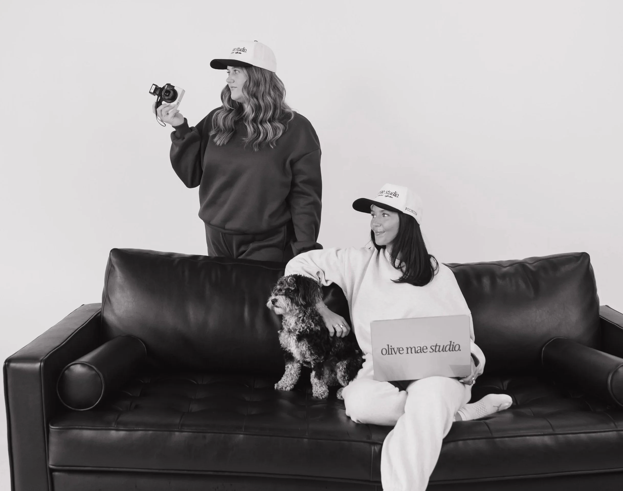 A woman with wavy hair wearing a cap and oversized sweatshirt, holding a camera. A young girl wearing a cap and hoodie, sitting on a black leather couch with a dog on her lap, wearing a bag with the text 'olive mae studio' on it. The scene is set against a plain white wall.