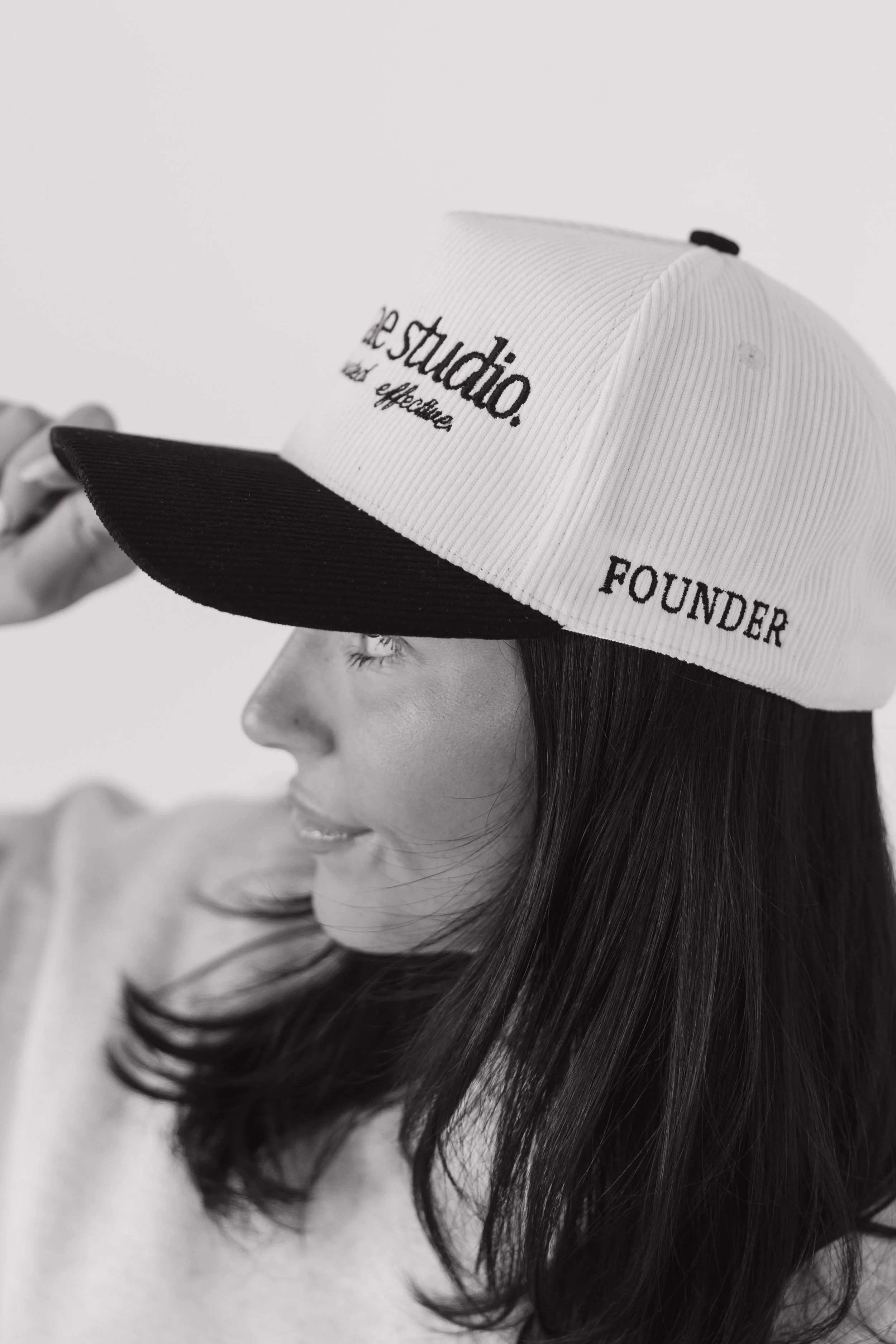 Black and white photo of a woman wearing a baseball cap with the words 'de studio?' and 'FOUNDER' embroidered on it, with her long dark hair flowing over her shoulder.