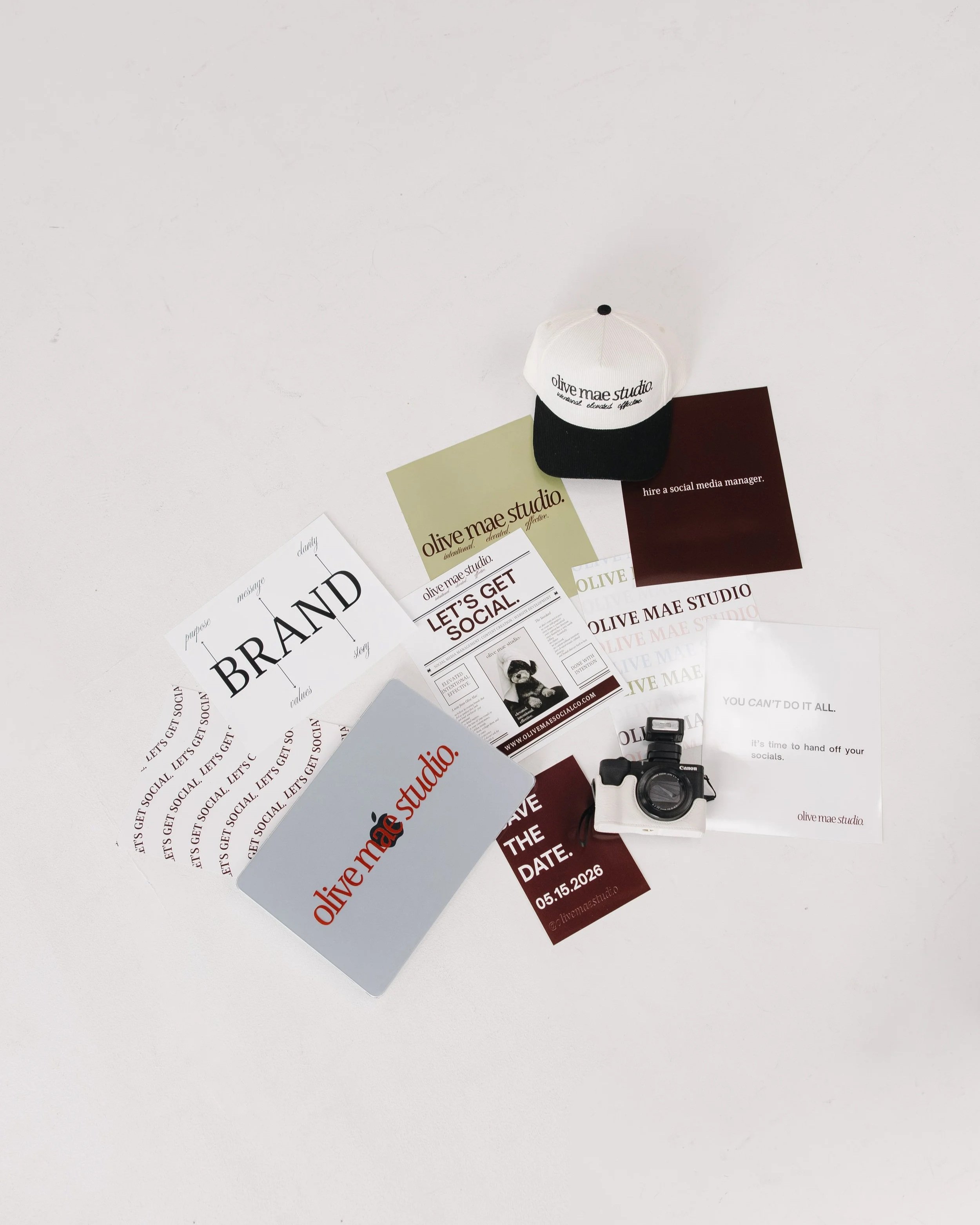Collection of printed flyers and cards with branding for Olive Mae Studio, a black and white camera, a white cap with black text, and a digital camera on a plain white surface.