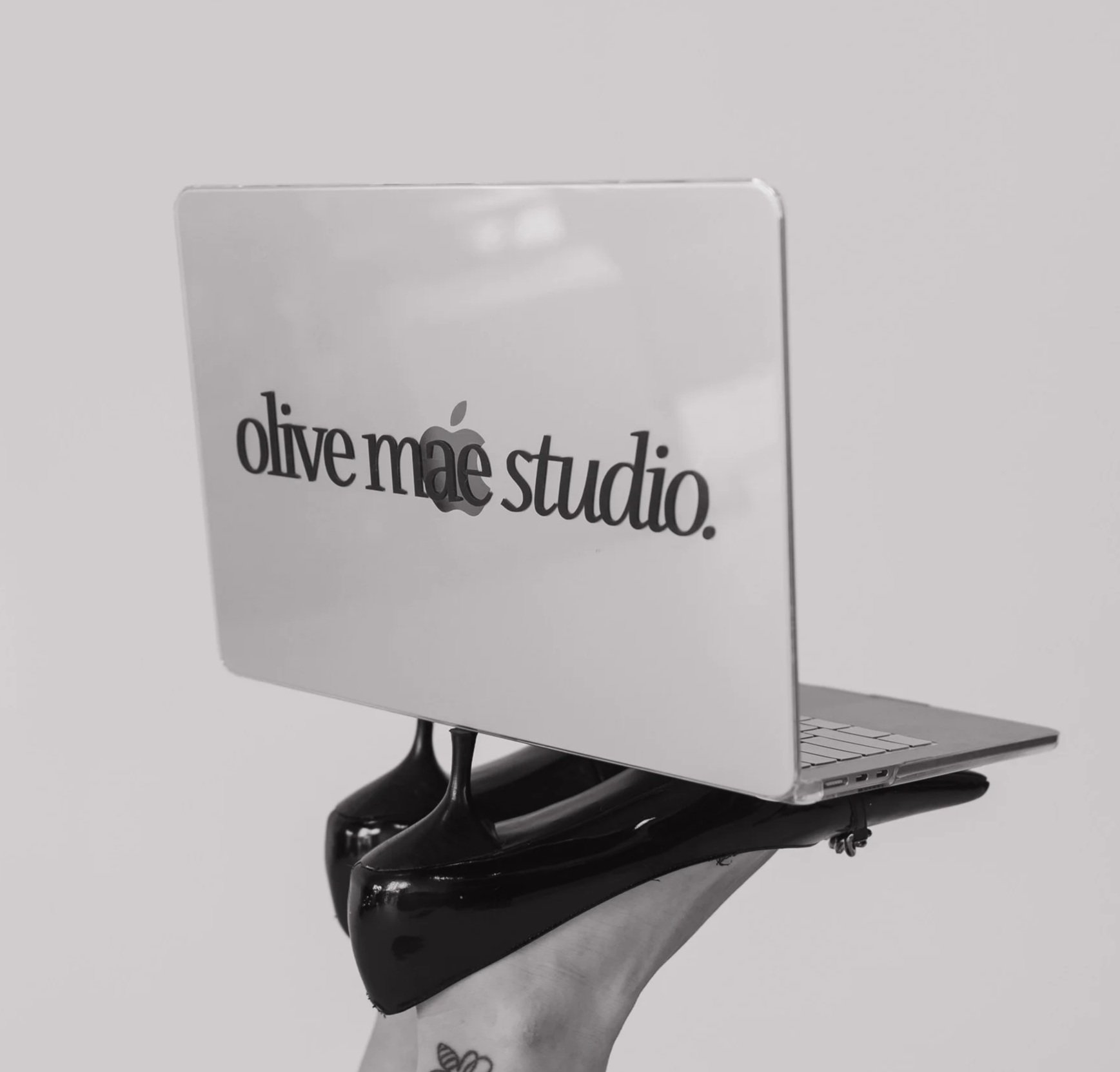 A person holding a skateboard with a miniature white MacBook in front of the skate, displaying a sign that reads 'olive máe studio.' in black text.