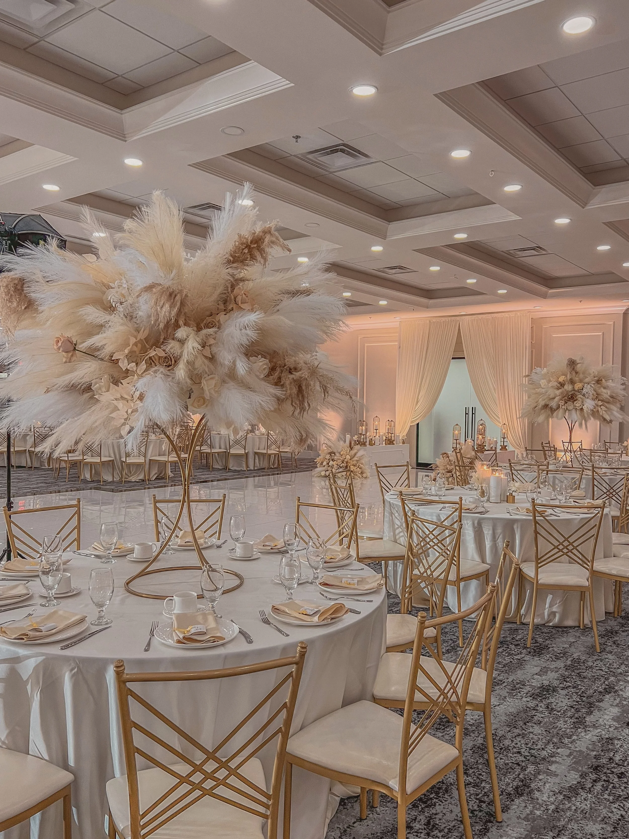 Elegant event hall decorated in soft neutral tones with round tables, gold chairs, and tall centerpieces of pampas grass and cream-colored flowers.