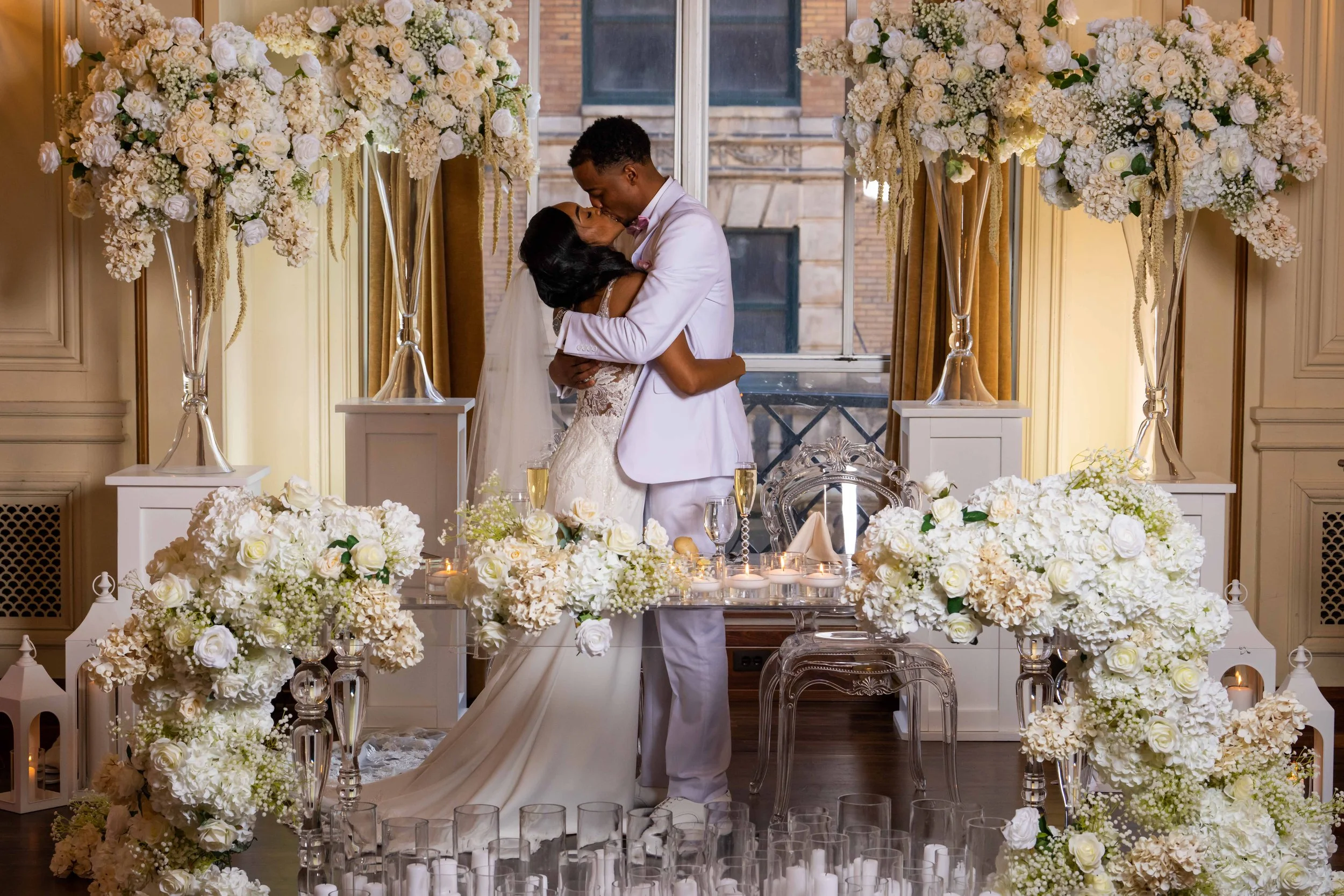 A newlywed couple sharing a kiss during their wedding ceremony, surrounded by large white floral arrangements and candles on a decorated altar.