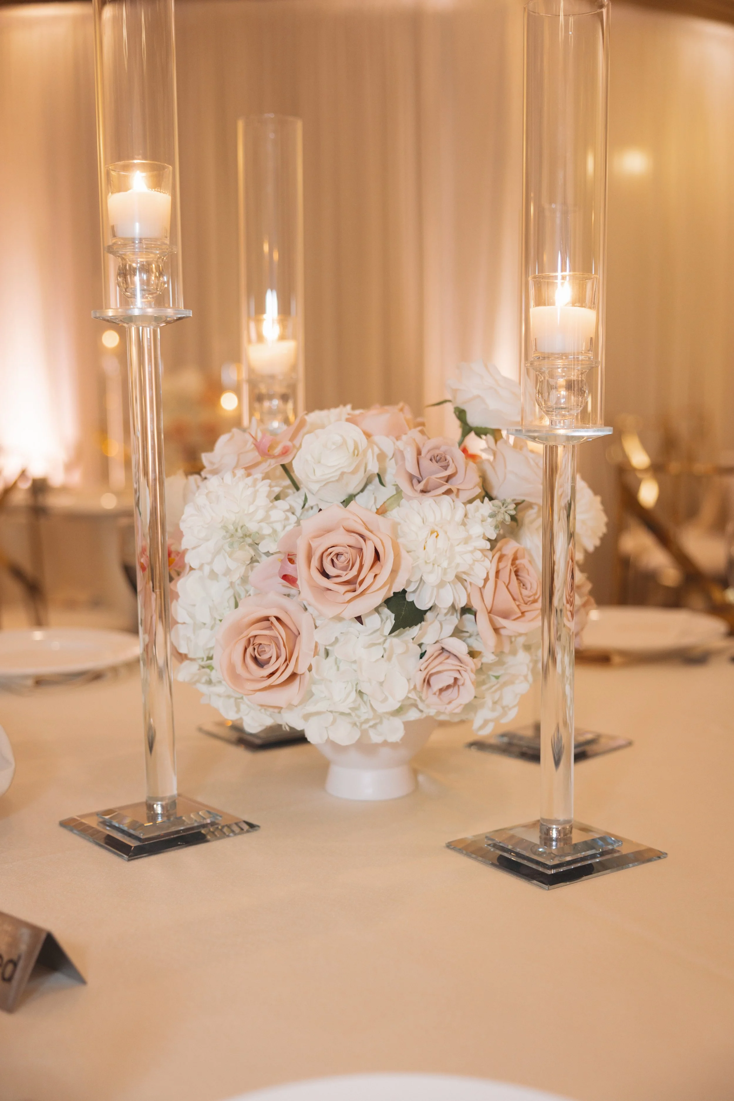 Elegant table centerpiece with a bouquet of pink and white roses and hydrangeas, surrounded by tall glass candleholders with lit white candles.