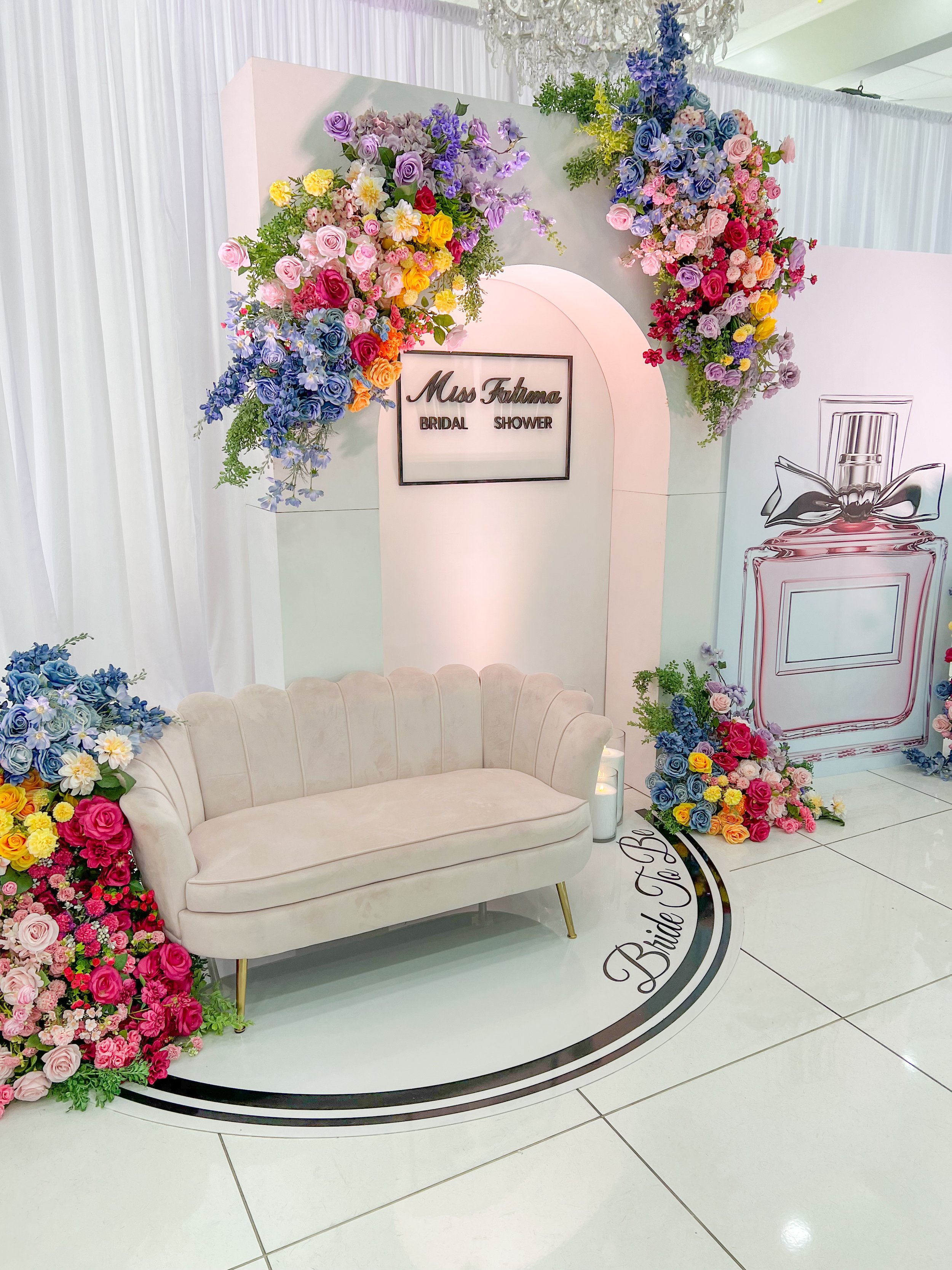 A decorated bridal shower setup with a white vintage-style sofa, surrounded by colorful floral arrangements of pink, purple, red, yellow, and blue flowers, and a white backdrop with a perfume illustration and a sign that reads 'Miss Futuma Bridal Shower'.