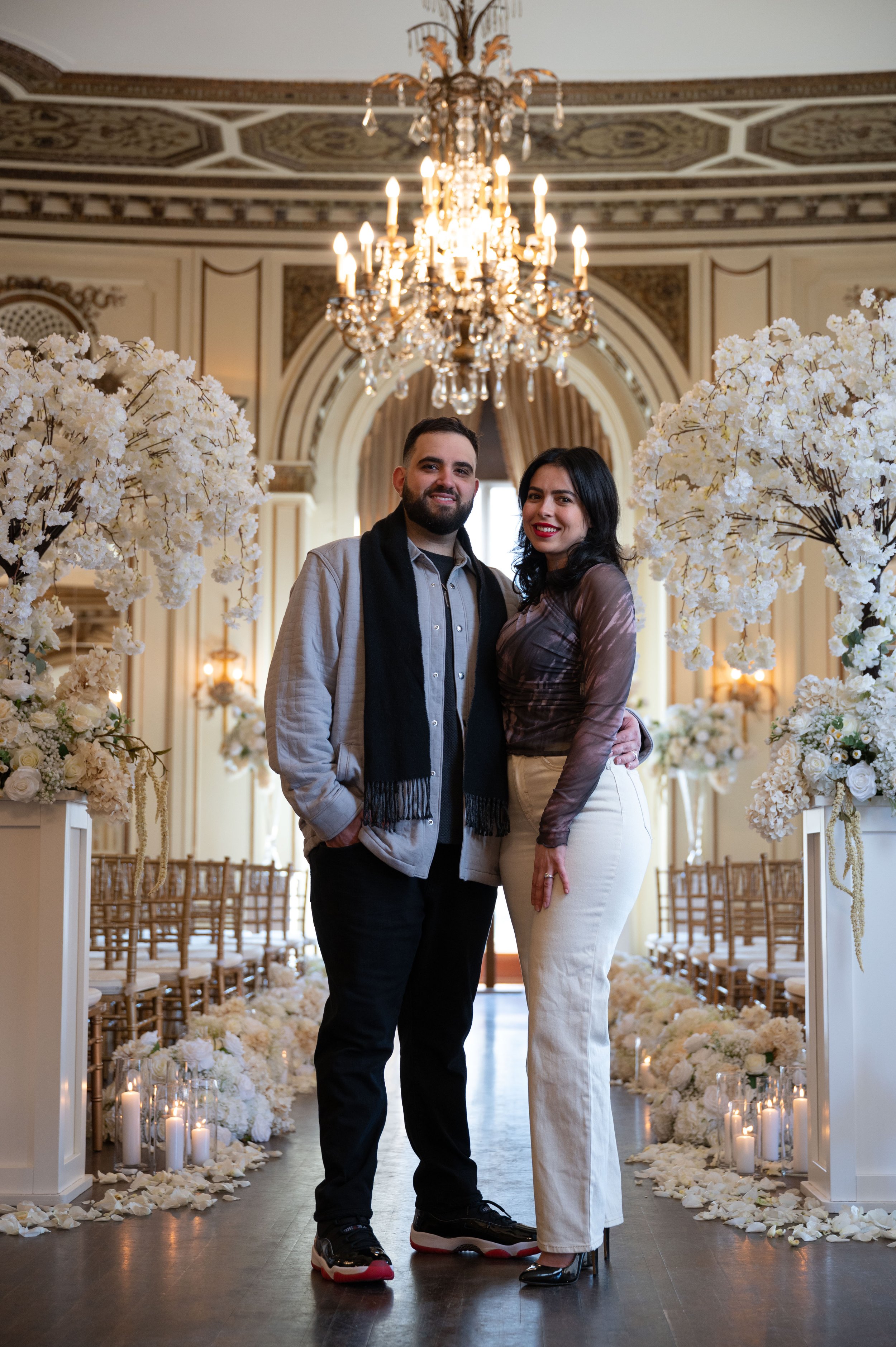 A couple standing in the center of a decorated wedding aisle with white flowers and candles, inside an ornate, chandelier-lit venue.