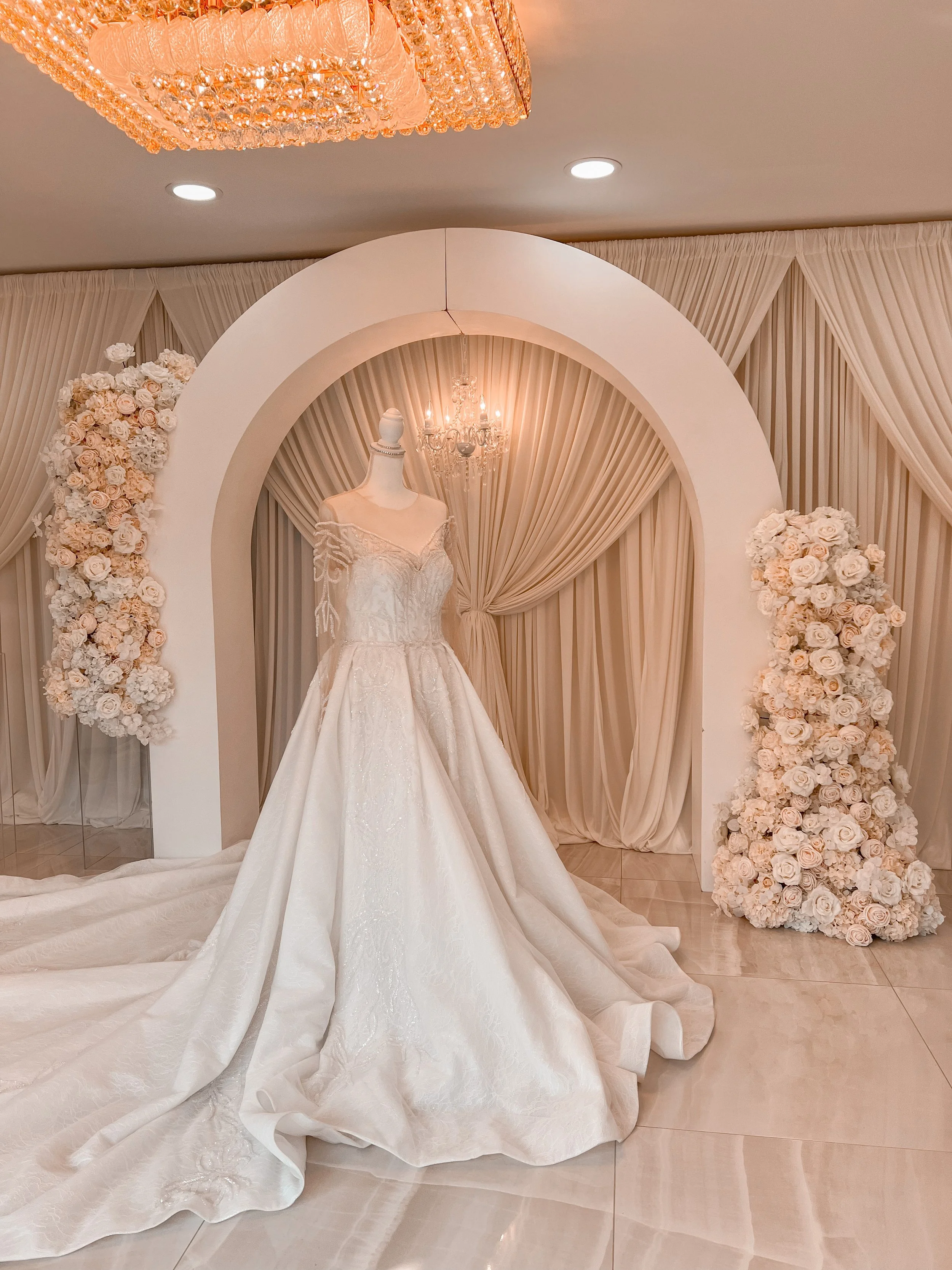 A wedding dress display on a mannequin, placed on a bridal stage with draped curtains and floral arrangements, illuminated by a chandelier above.