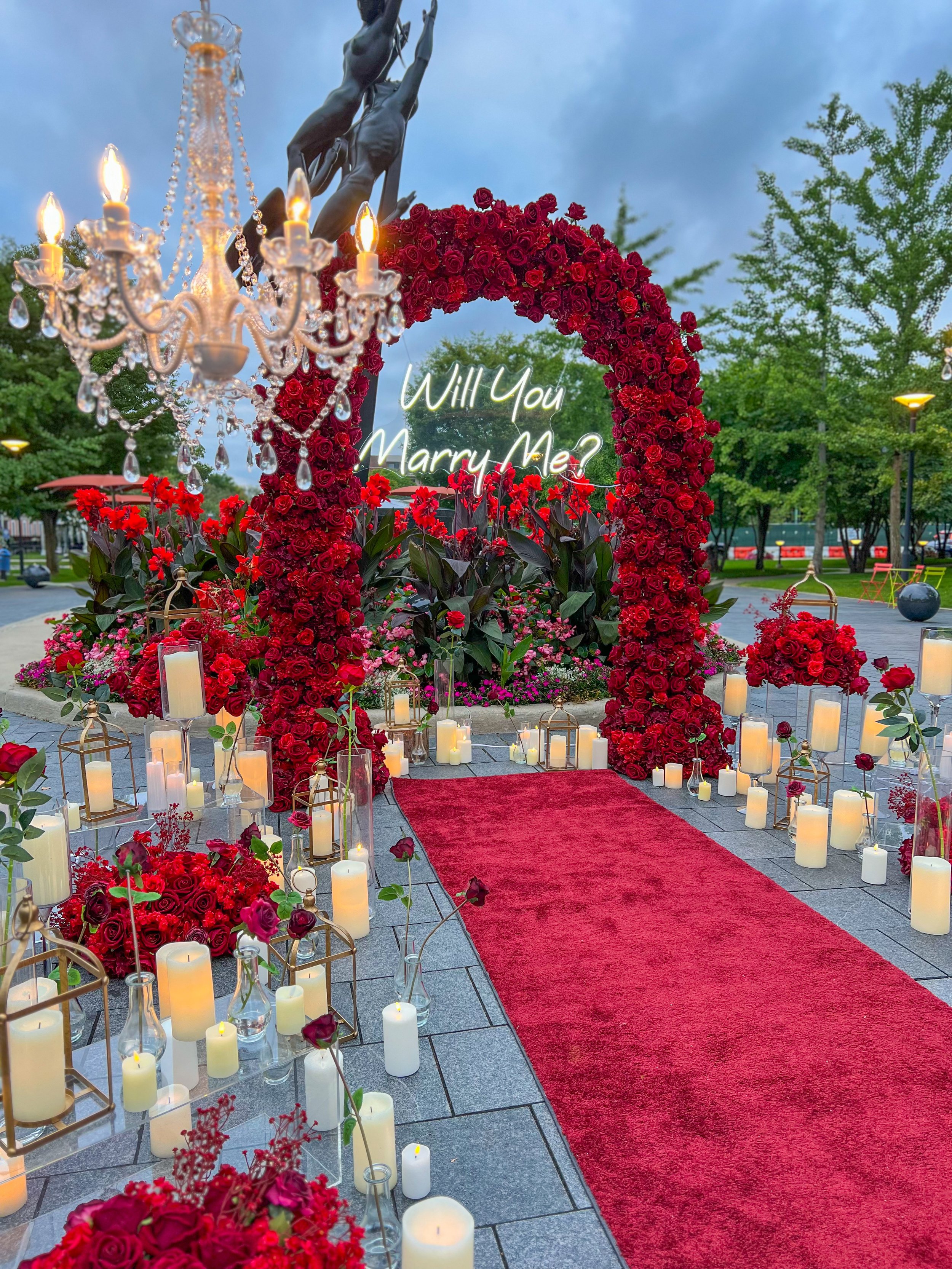 A romantic outdoor scene with a red carpet leading to a floral arch made of red roses. Candles in glass and lantern holders are arranged along the carpet edges or on the ground. A neon sign inside the arch says, 'Will You Marry Me?' A chandelier hangs from above, and in the background, there are trees and a sky with clouds.