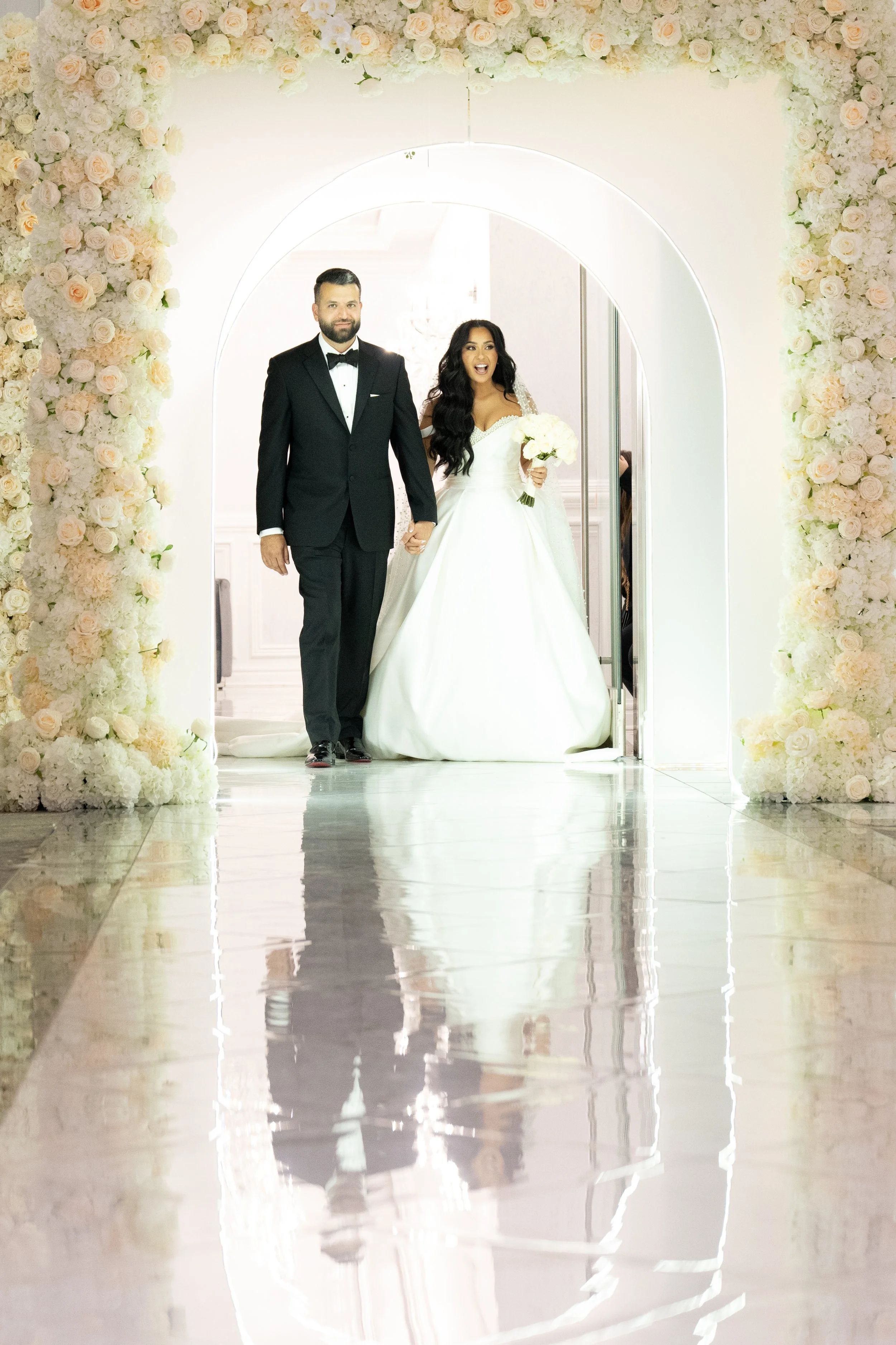 A bride and groom walking through a flower-decorated archway at their wedding ceremony, holding hands and smiling. The bride is wearing a white wedding gown and holding a bouquet, while the groom is in a black tuxedo.