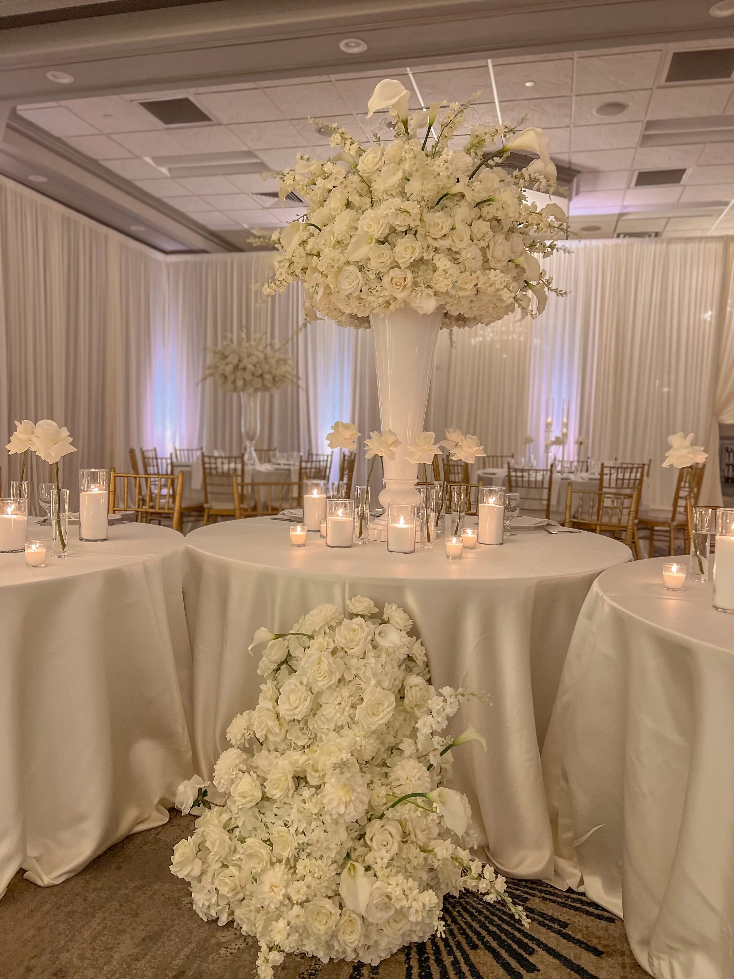 Elegant wedding reception table decorated with a tall white floral centerpiece, smaller floral arrangements, and lit candles with a backdrop of white drapes and ambient lighting.