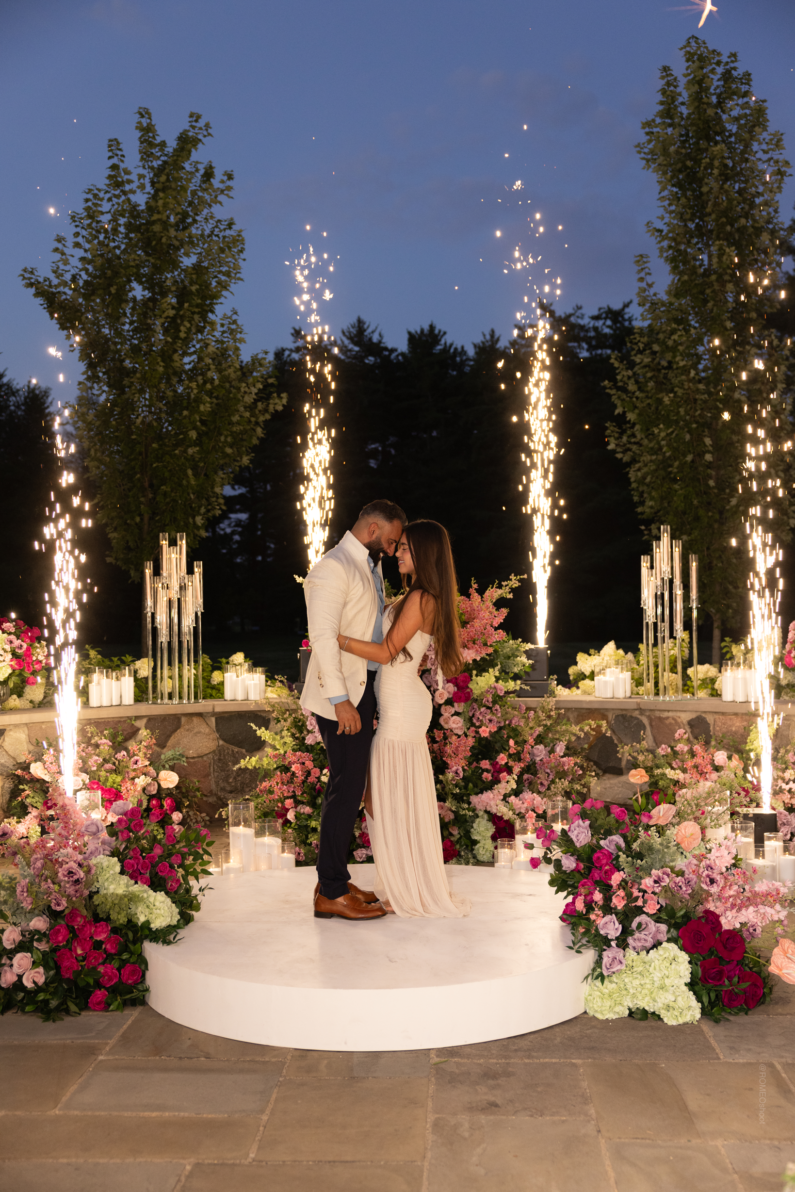 A couple in formal attire standing on a decorated white circular platform outdoors during evening, surrounded by pink and purple flowers, candles, and fireworks displays in the background.