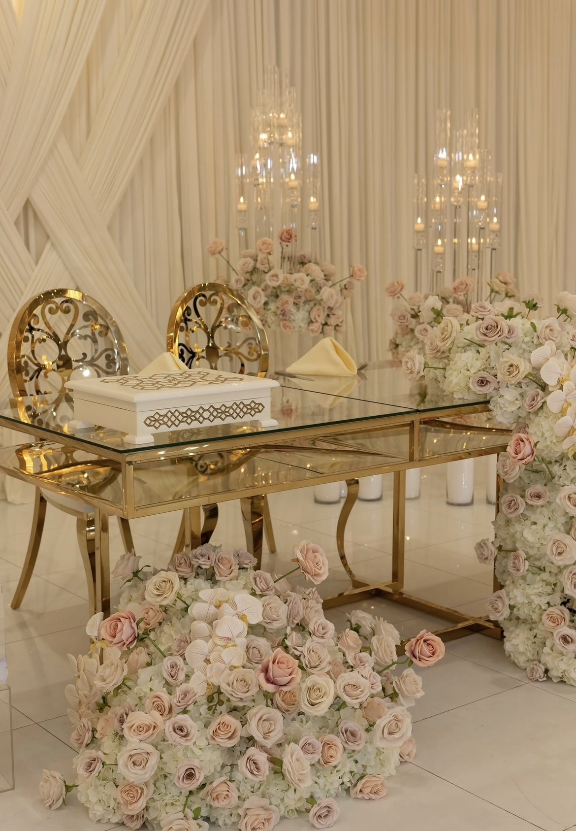 Elegant wedding reception table with gold accents, surrounded by floral arrangements of light pink and white roses, and topped with candles and chandeliers in the background.
