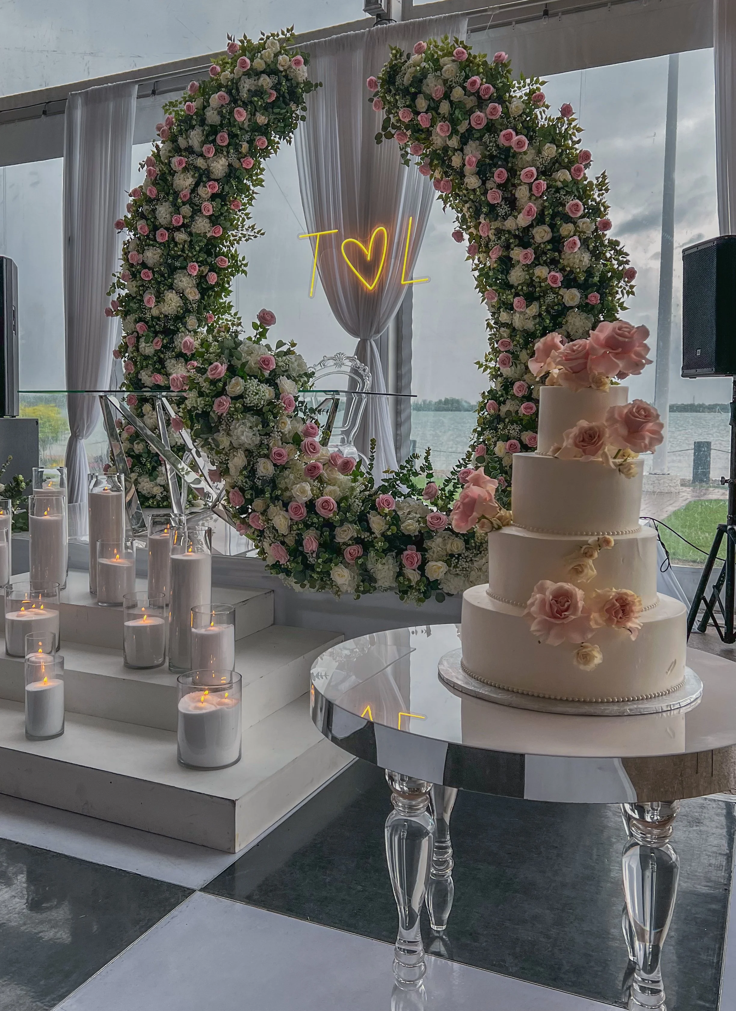 A three-tier white wedding cake decorated with pink and white flowers on a glass table, a large floral heart-shaped arch with pink and white roses, and a background view of water through windows.