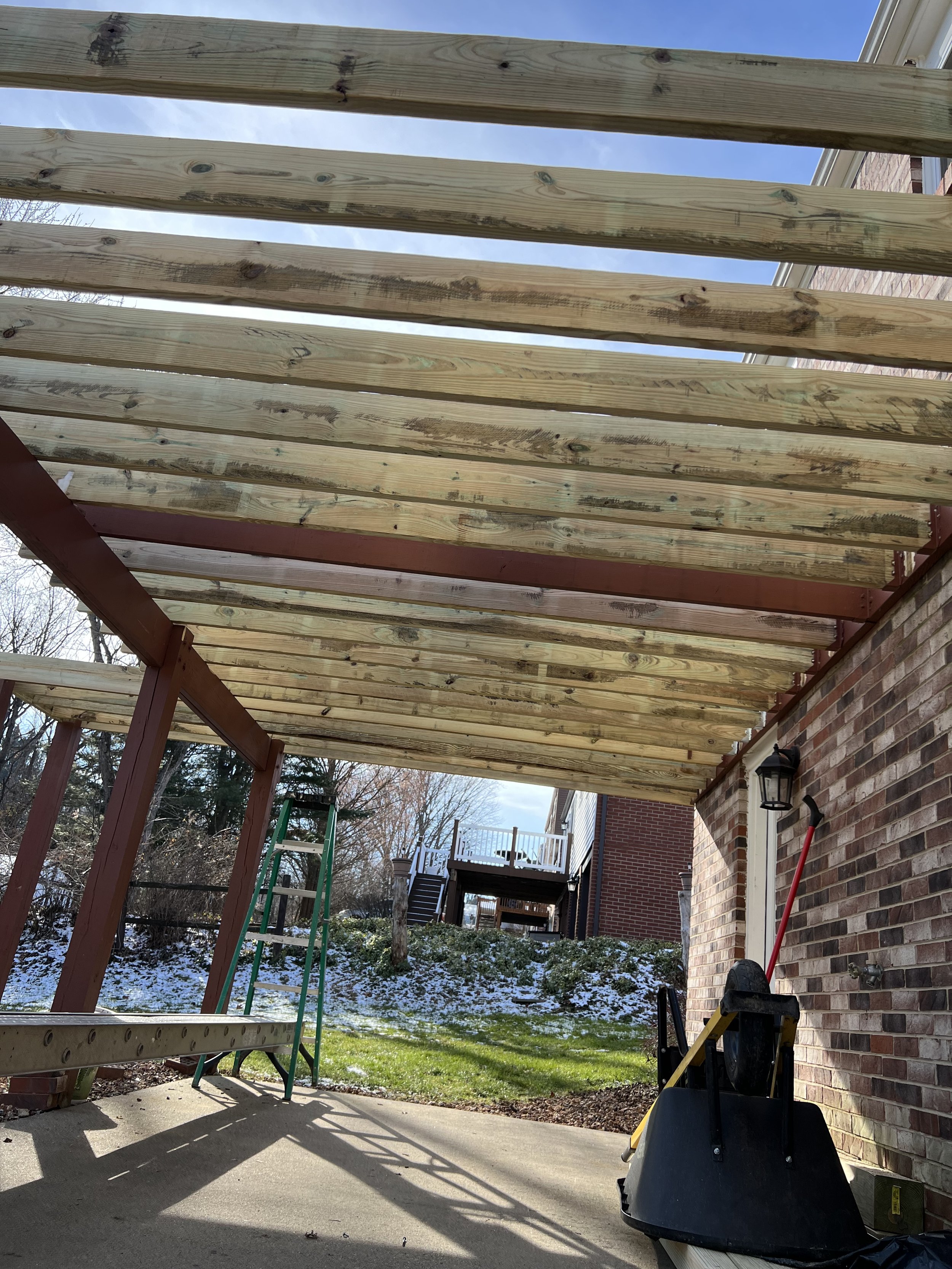Under construction patio cover with wooden beams, a ladder, and yard tools, in a backyard with snow patches, trees, and neighboring houses.