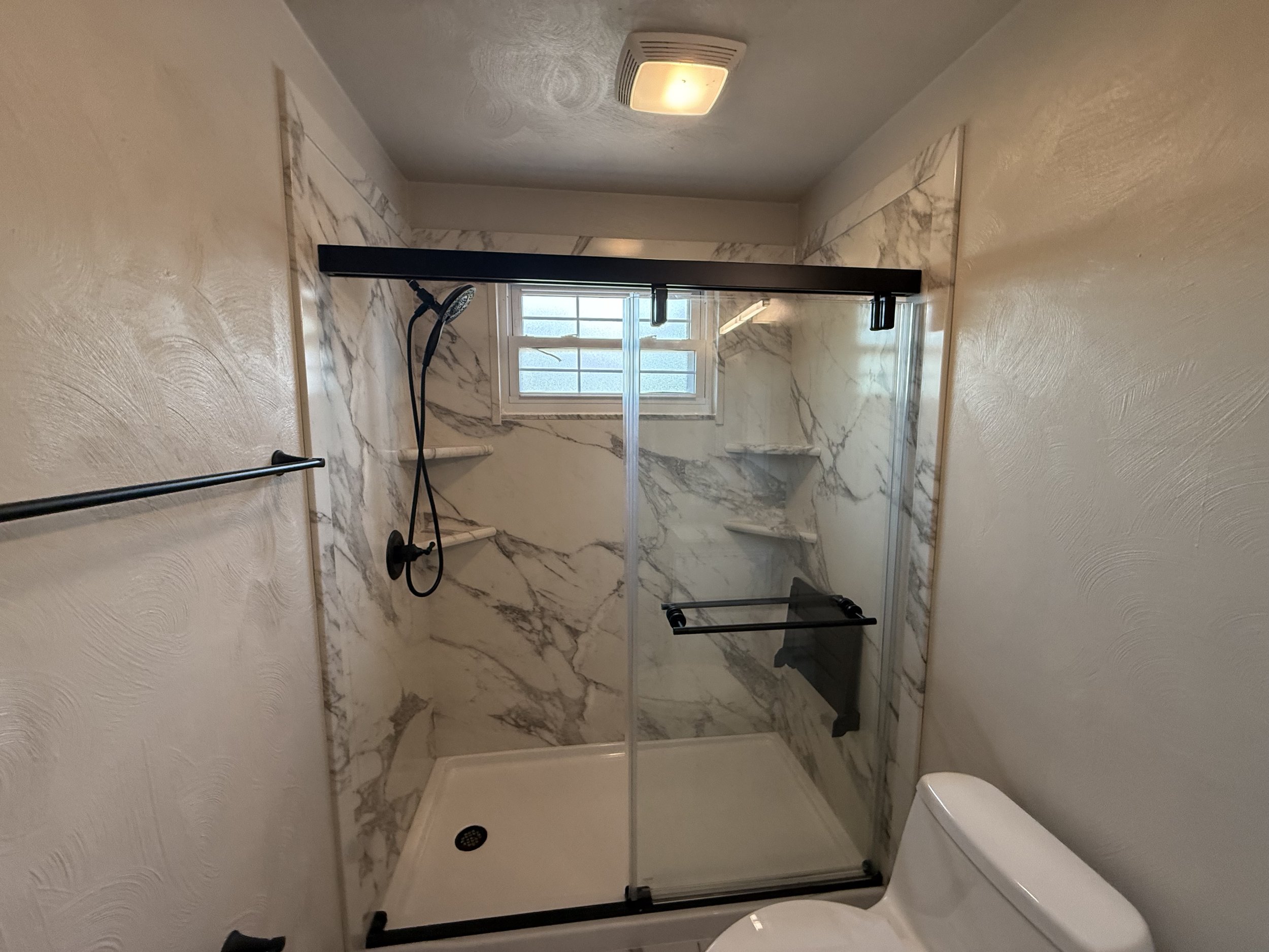 Small bathroom with marble shower enclosure, black framed glass door, wall-mounted showerhead, white toilet, beige textured walls, and small window.