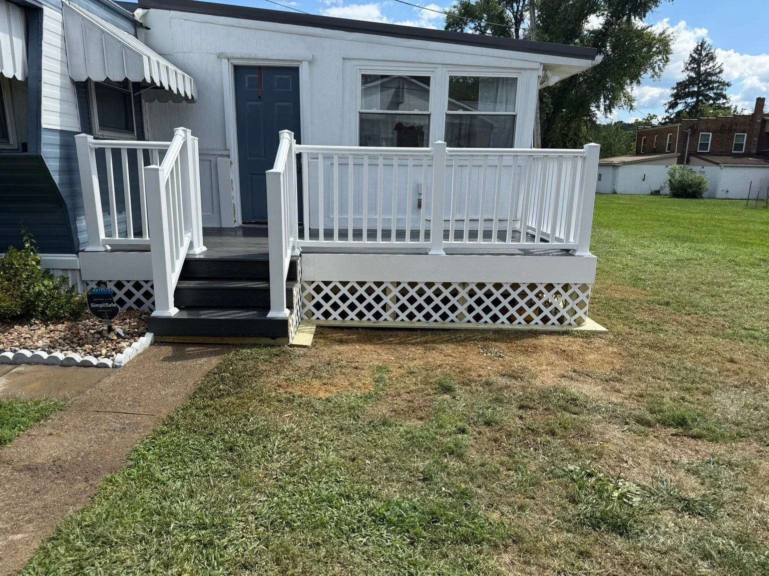 White wooden deck attached to a small house with steps leading up to it, featuring white railings, and a sliding door, surrounded by a grassy yard.