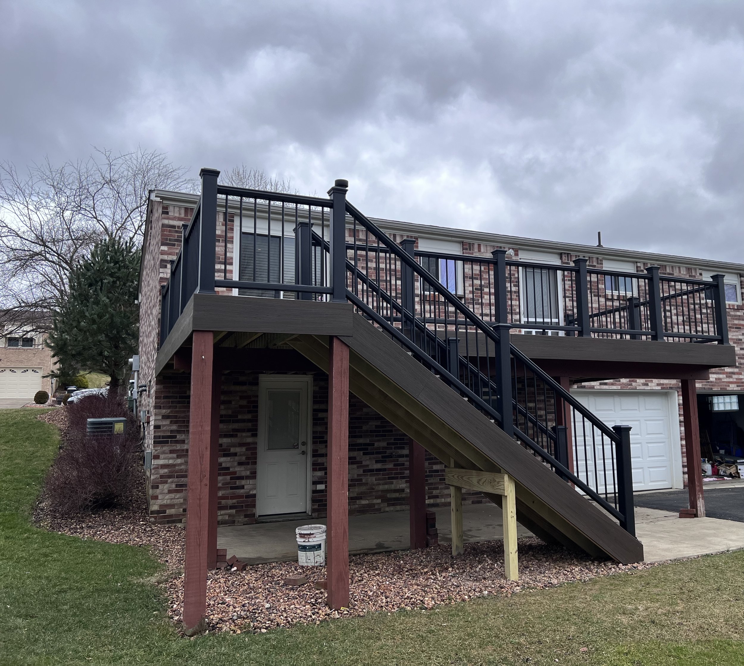 The image shows a two-story brick house with an outdoor staircase leading to a second-story deck with black railings, under overcast skies.