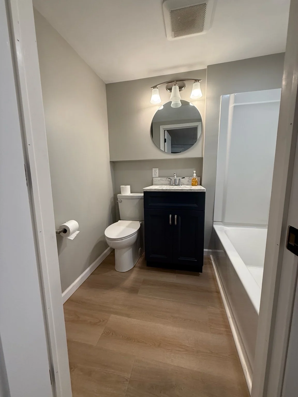 A modern bathroom with a toilet, a dark vanity with a marble countertop, a round mirror, a wall-mounted light fixture, a sink with a soap dispenser, and a bathtub with a white surround. The bathroom has beige flooring and light-colored walls.
