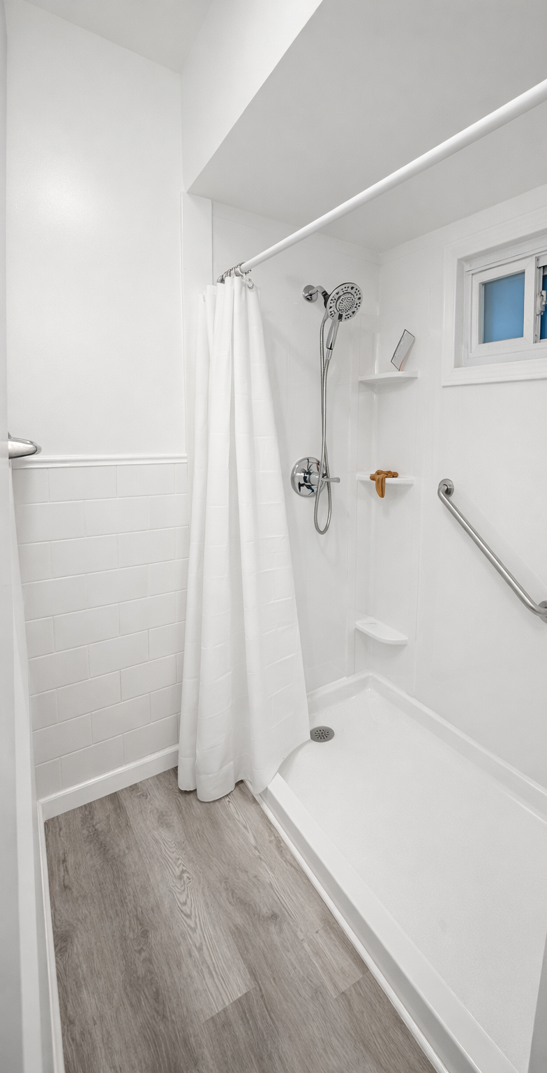Completed residential bathroom remodeling project in Vandergrift, Pennsylvania. This image showcases a renovated tub and shower featuring a new white acrylic surround, updated plumbing fixtures, integrated grab bars, luxury vinyl plank flooring, and 