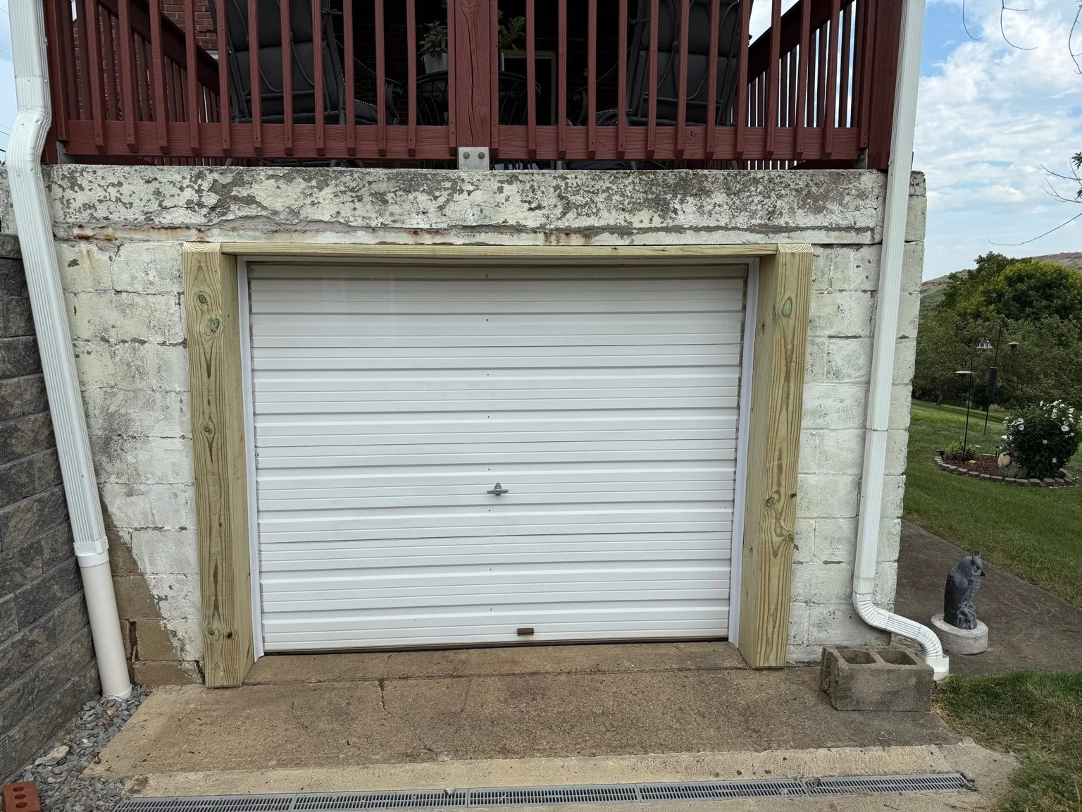 A white garage door with a small lock, surrounded by new light-colored wooden framing, set into a concrete and brick wall. Above is a wooden deck with red railing, and to the right is a landscaped yard with a statue of a dog, green grass, and trees.
