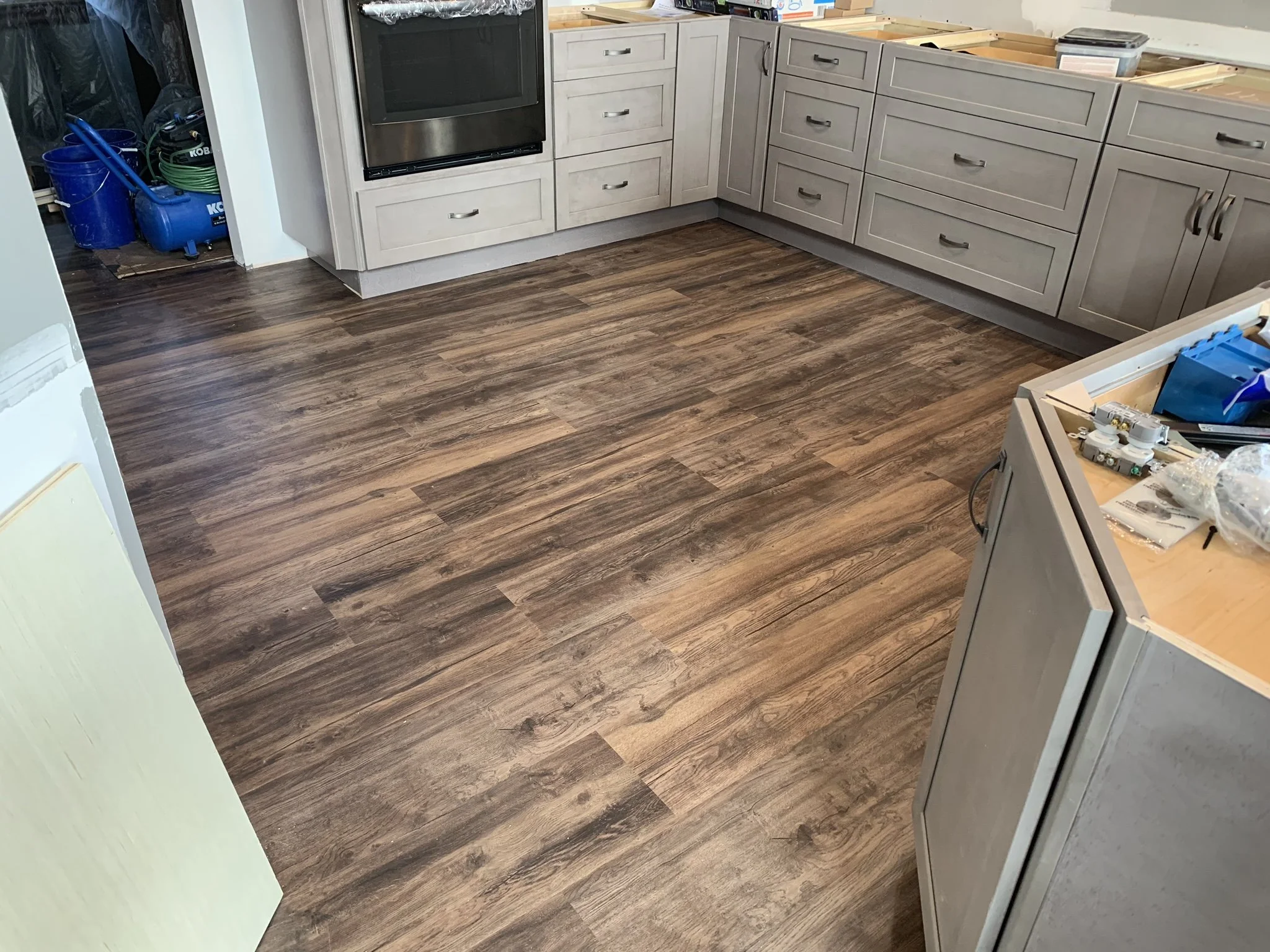 Kitchen with wood-look flooring, gray cabinets, and stainless steel oven. Construction tools and materials are visible on some countertops and floor.
