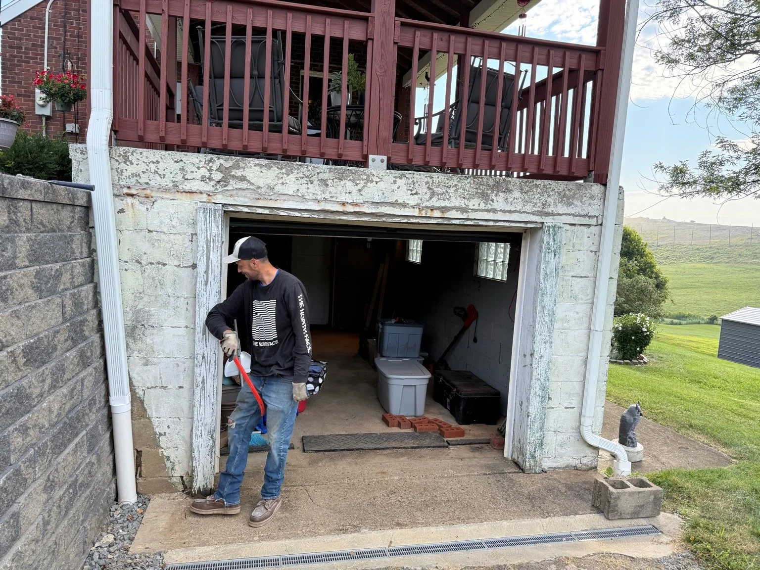 A man working outside a garage with a red-handled tool, in a residential backyard with a deck above, some garden tools, storage containers, and a grassy area with rolling hills in the background.