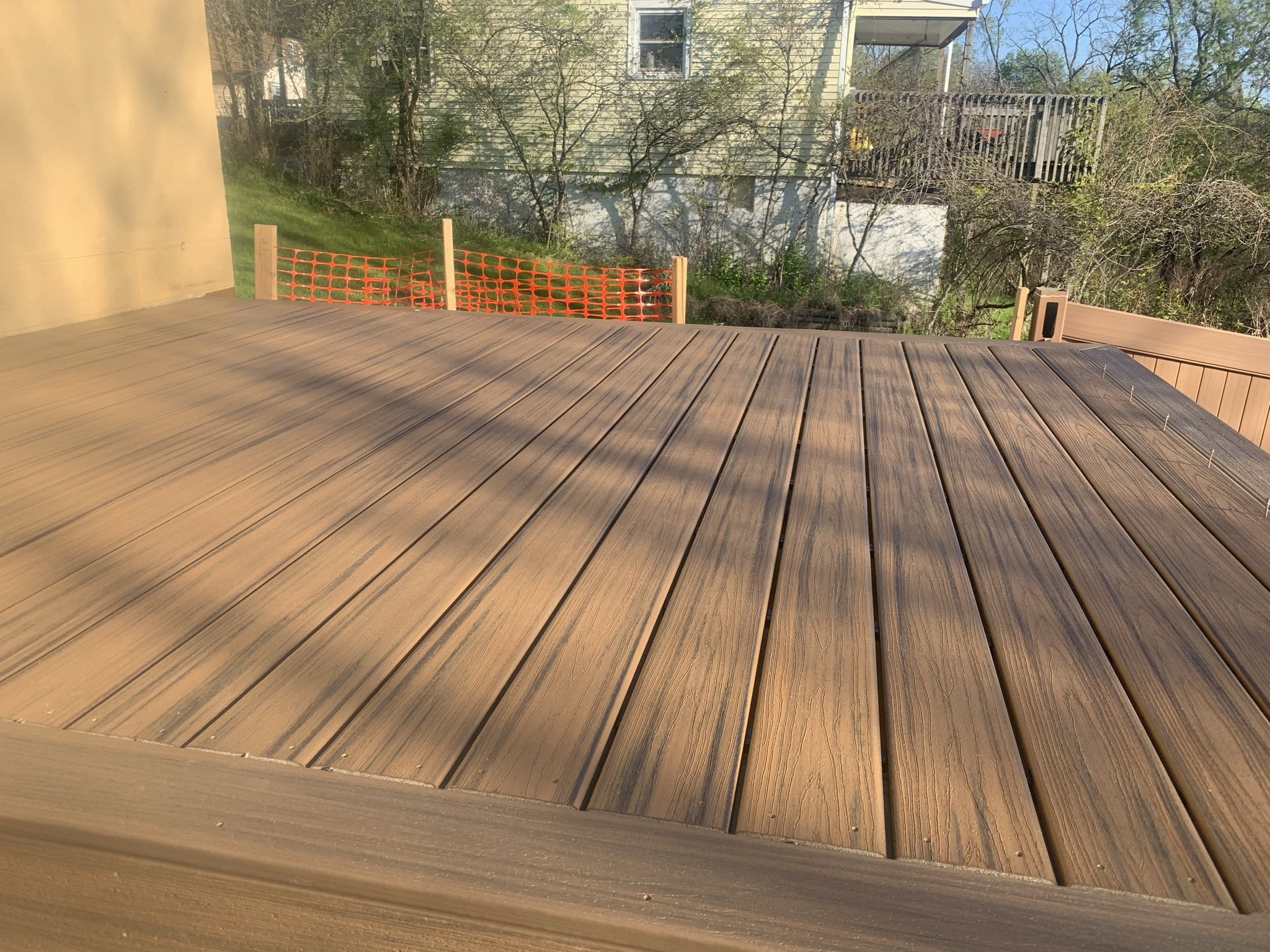 A newly installed wooden deck with light brown planks, surrounded by a safety orange plastic fence, in a suburban backyard with trees and neighboring houses in the background.