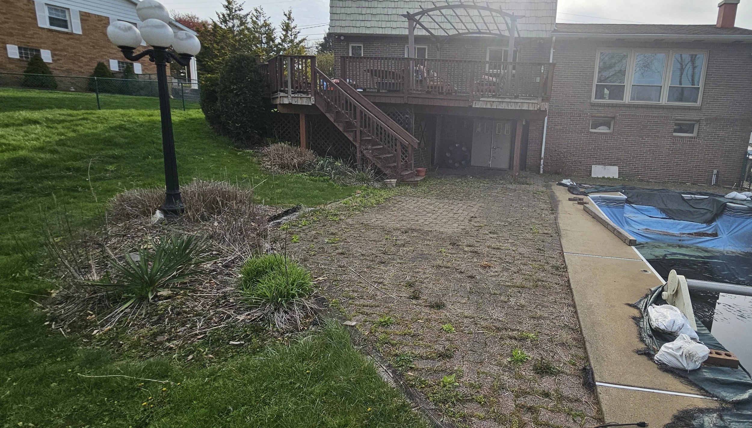 Backyard with a patio, a raised deck with stairs, a pond under construction with a liner, and a street lamp among shrubs and grass.