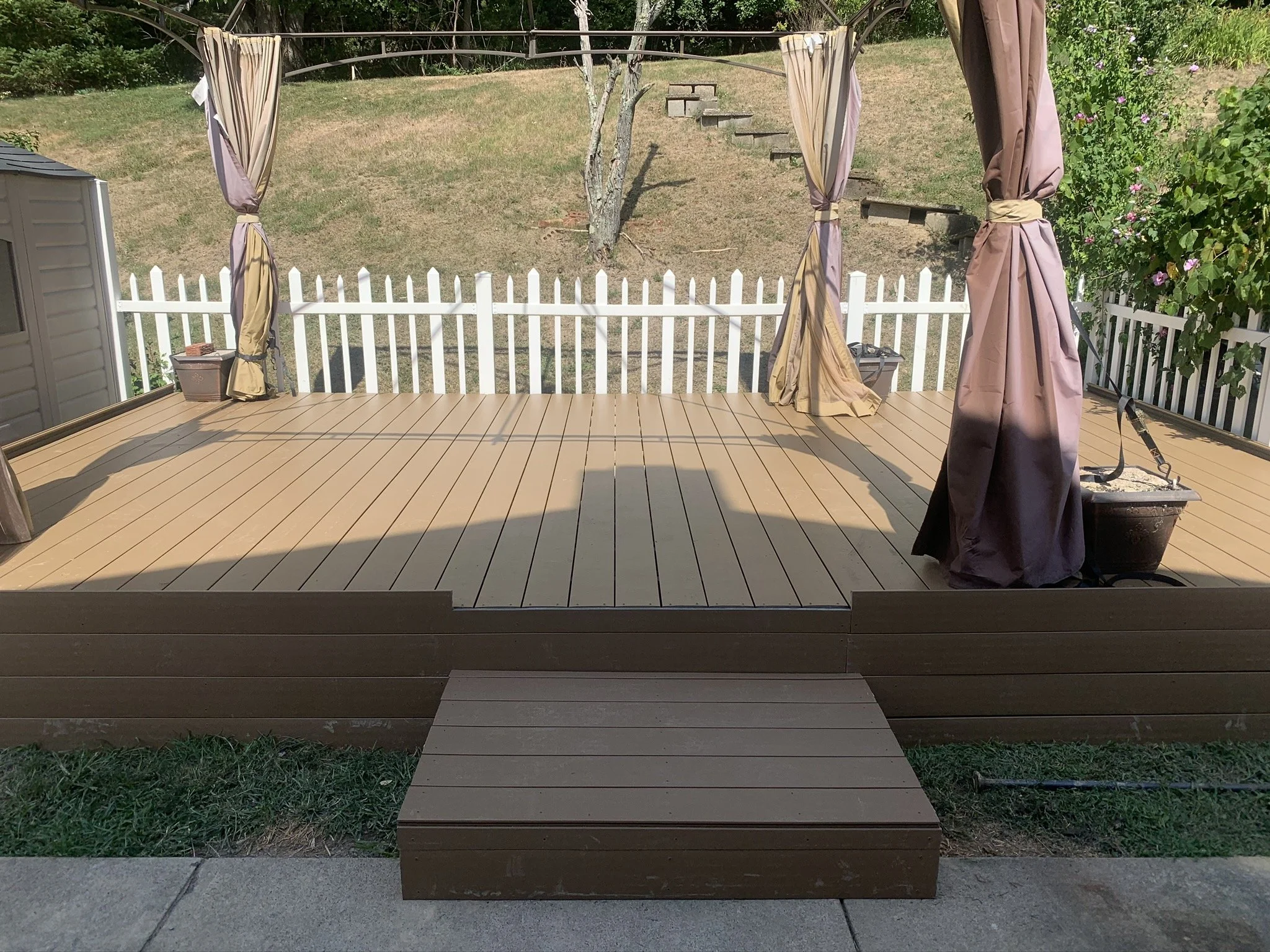 A backyard deck with brown wooden planks, bordered by a white picket fence, and decorated with brown and beige curtains tied back to posts. There is a set of steps leading up to the deck, and some gardening tools and planters are visible.