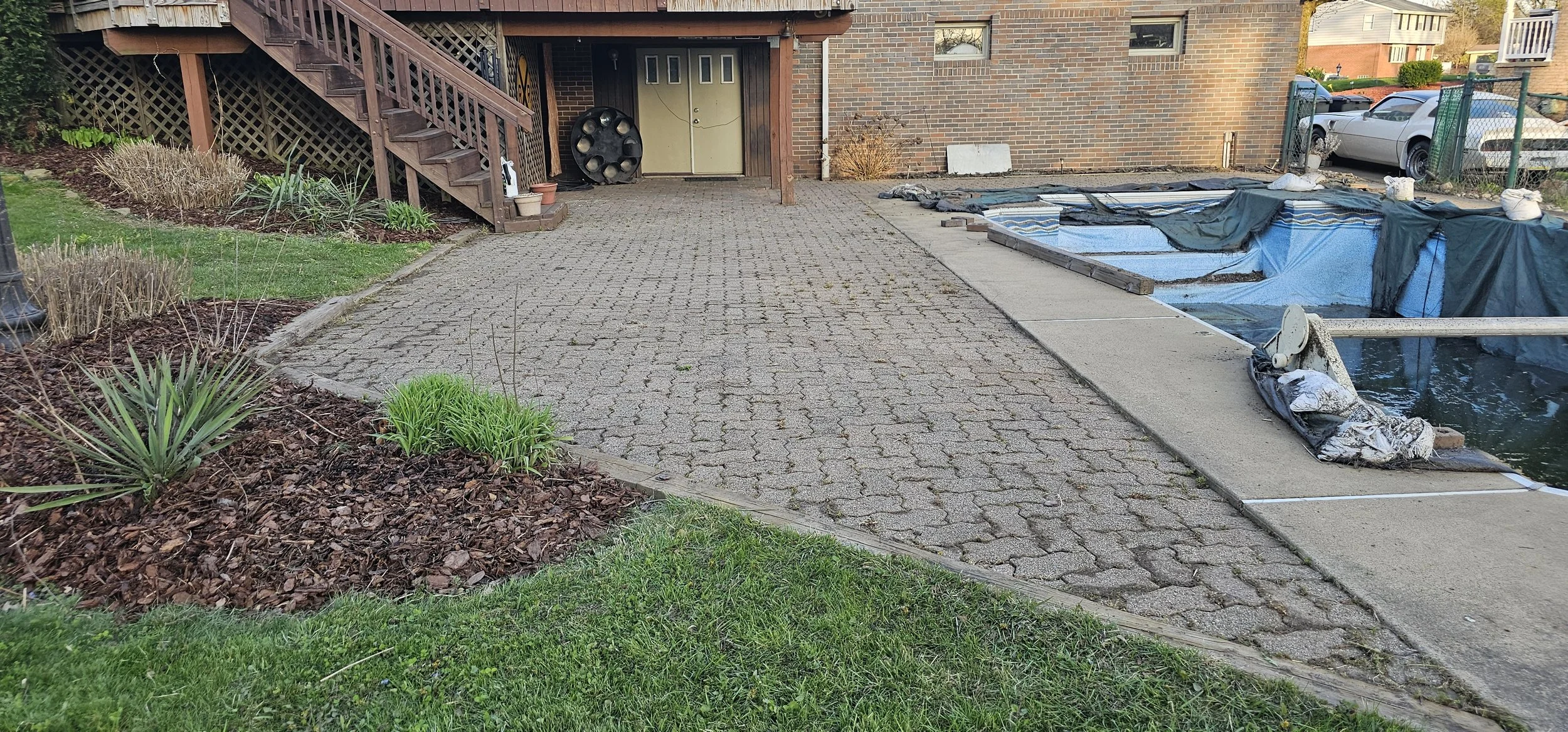 Residential backyard with a brick and wood house, showing a paved patio, garden beds with plants, a staircase leading to a deck, and an in-ground swimming pool under construction with blue tarp and tools around.