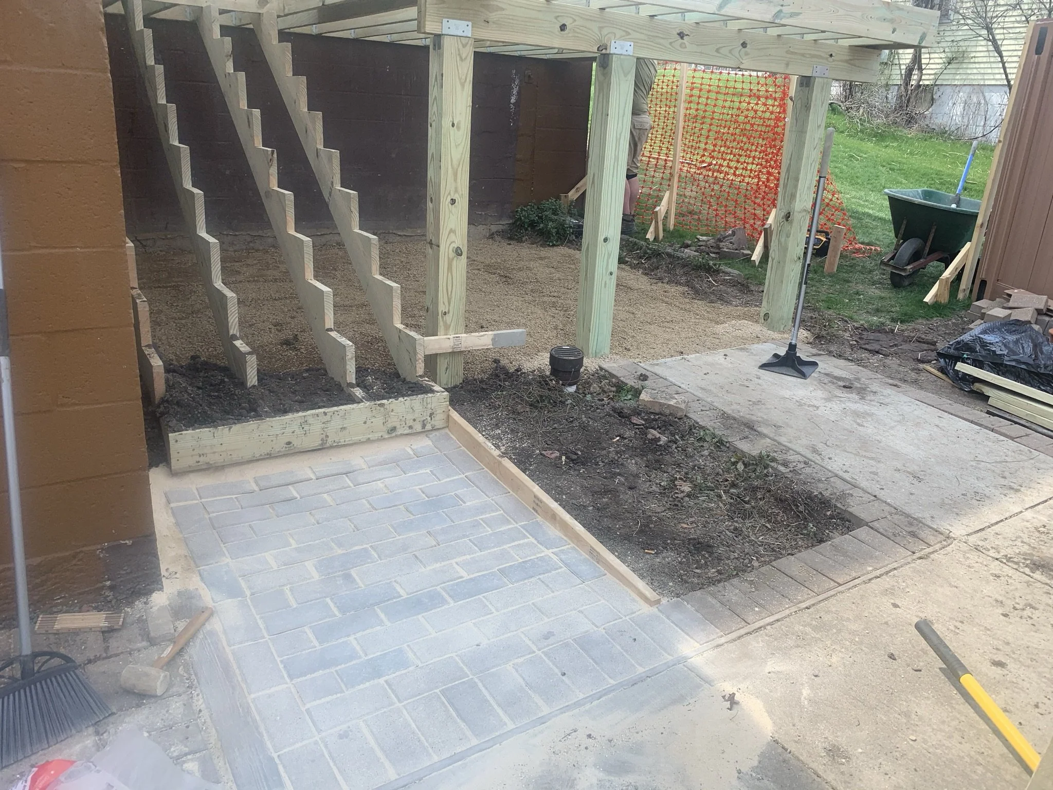 Under construction patio with newly laid gray brick pavers, a wooden staircase frame, and garden tools like rake and hammer nearby.