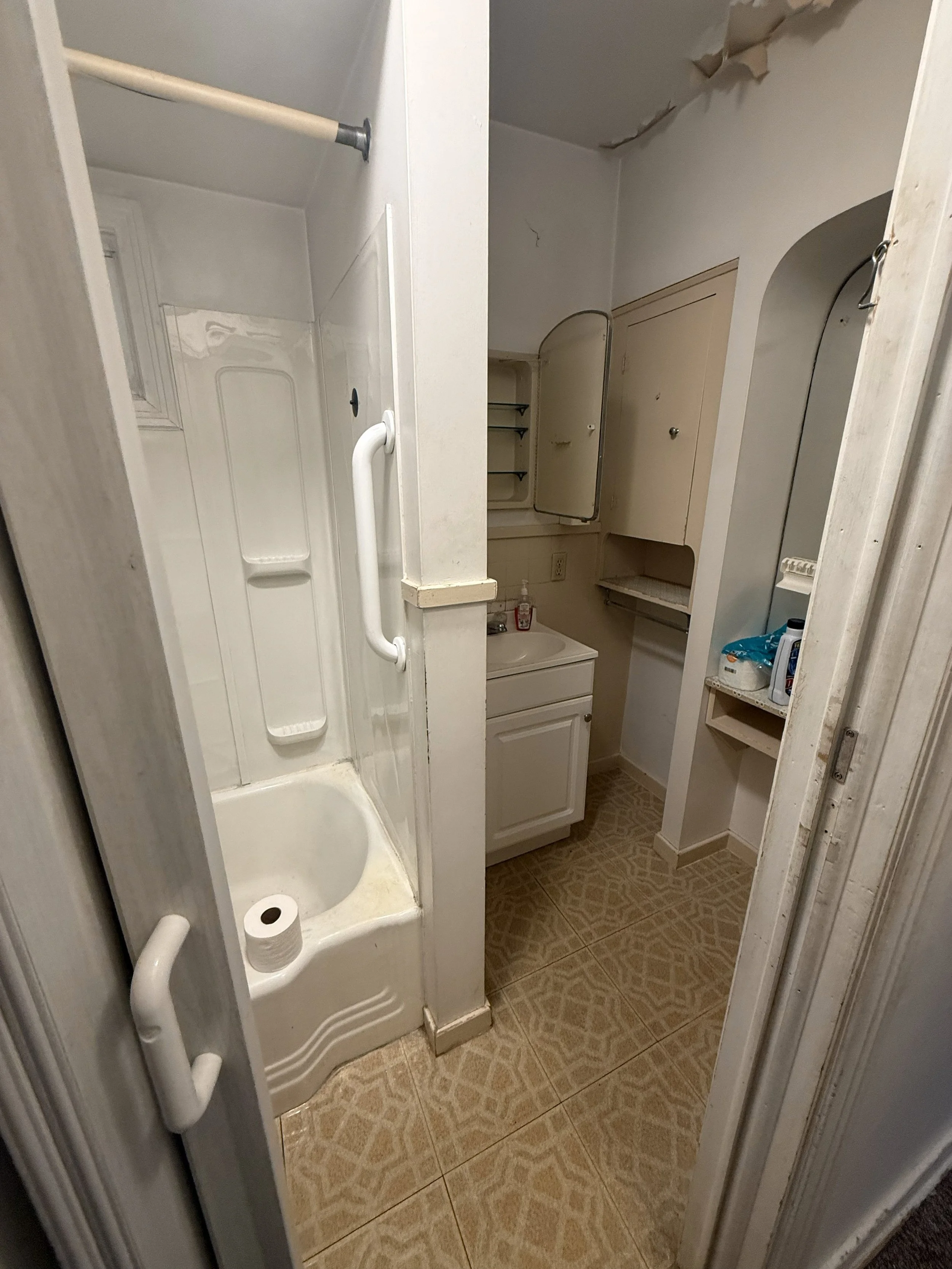 Before photo of a residential bathroom remodeling project in Vandergrift, Pennsylvania. This image shows the original bathroom featuring a fiberglass tub and shower, compact vanity, original toilet, vinyl flooring, and existing finishes prior to reno