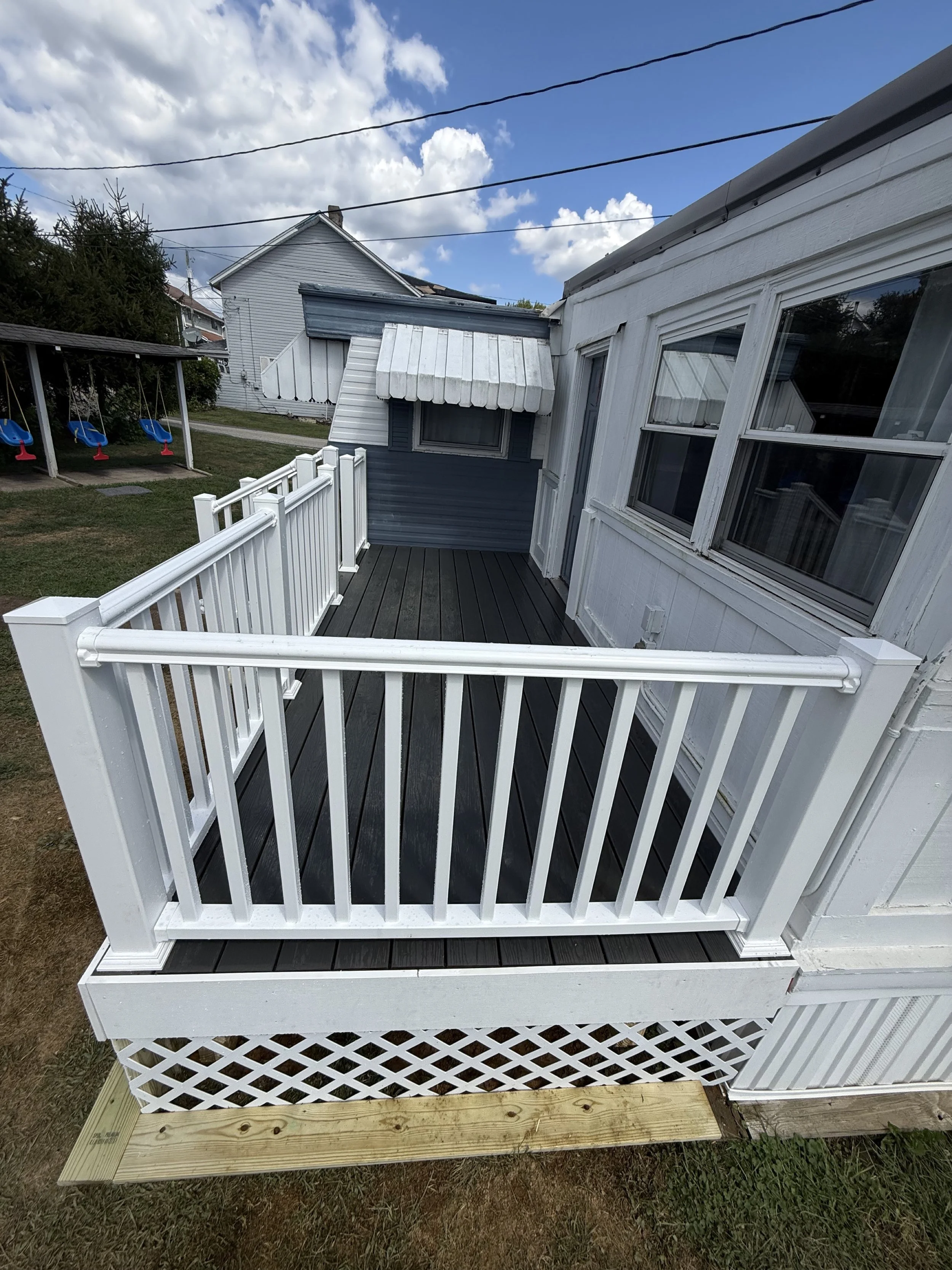 Small outdoor deck with white railing, attached to a house with white siding. Behind the deck, there is a small blue building with a window and a white awning. The yard has grass, and in the background, there are houses, a swing set, and a partly clo