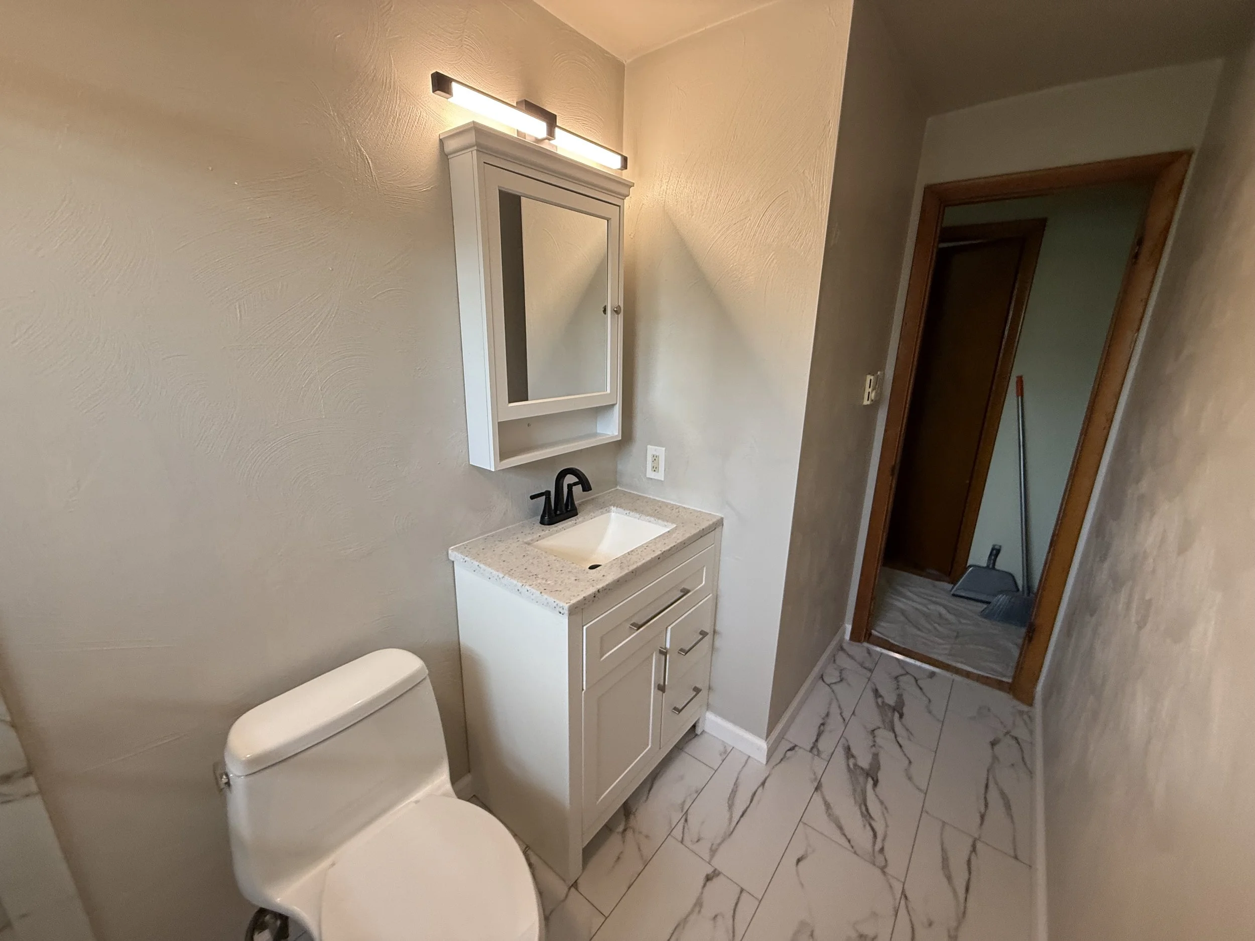 Small bathroom with a white toilet, a white vanity with a black faucet, a mirrored medicine cabinet, wall-mounted light fixture, and a doorway leading to a laundry area with a broom and dustpan on the floor.
