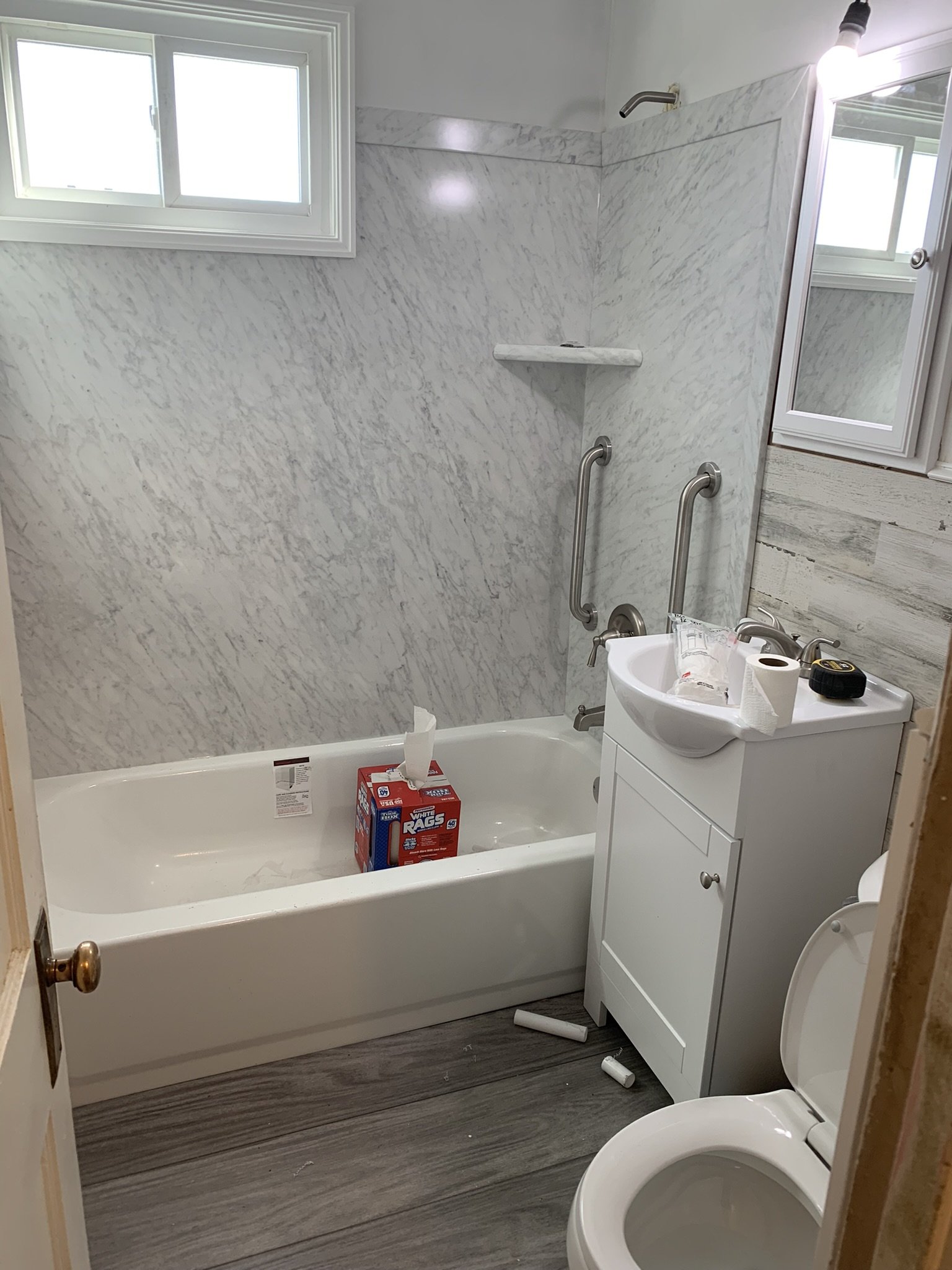 A small bathroom with a bathtub, a toilet, a sink cabinet, and two small windows. The wall tiles are light gray with a marble pattern. The bathtub has a box of plastic trash bags inside. There's a roll of toilet paper and some small items on the sink