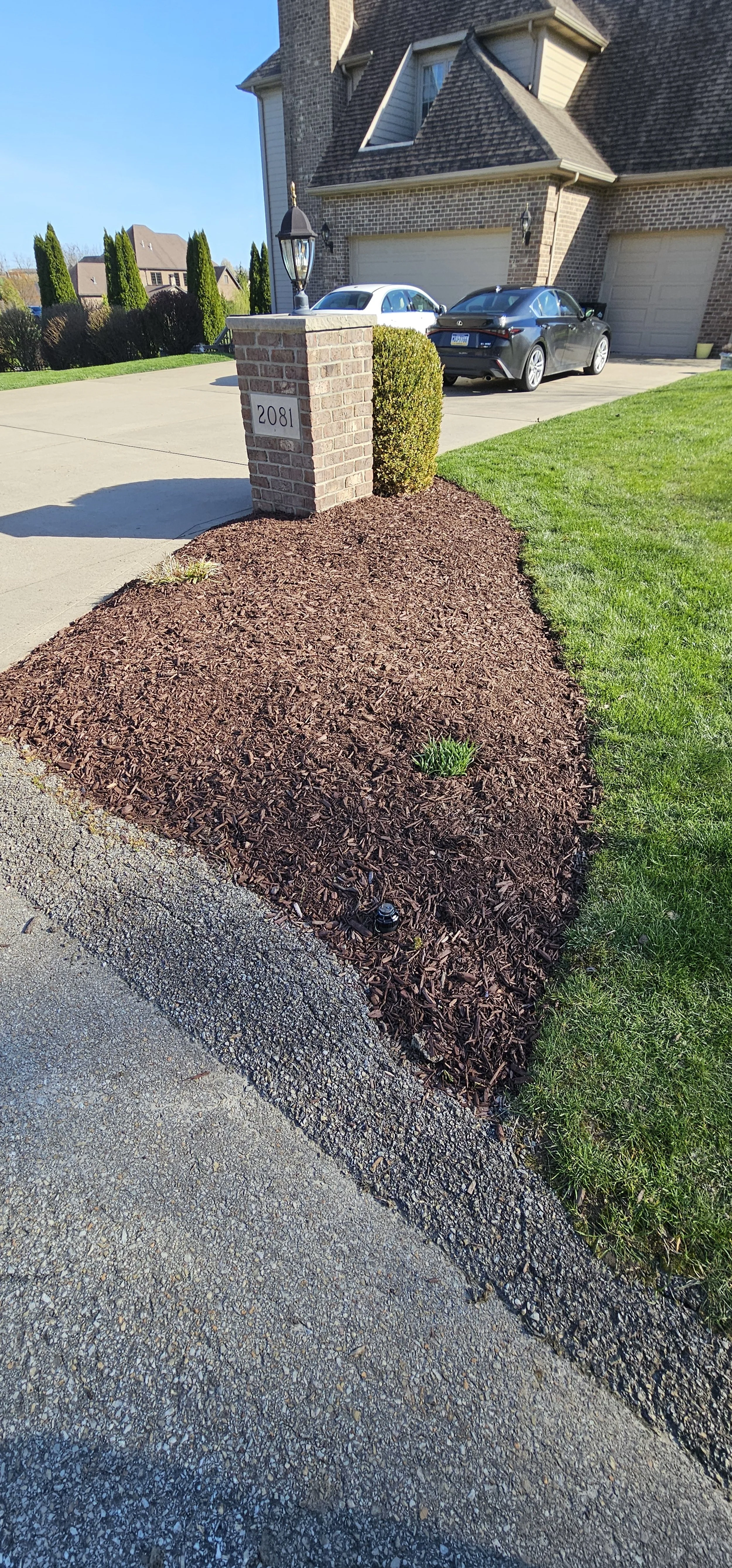A residential front yard with a brick mailbox, manicured shrub, and newly mulched flower bed next to a concrete driveway and house with two parked cars.