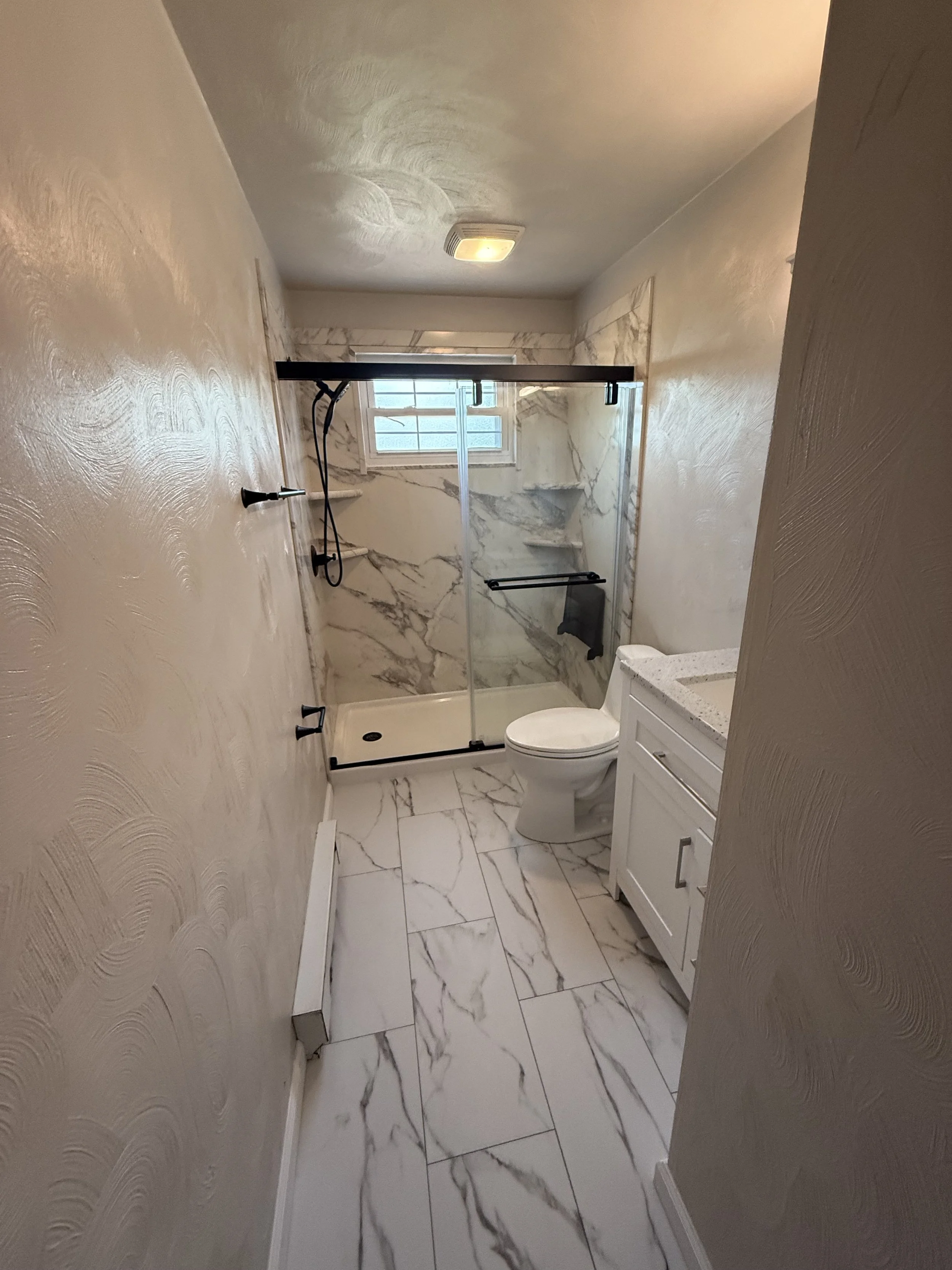 A small bathroom with marble tile flooring and marble wall in the shower area. It includes a glass-enclosed walk-in shower with a window, a white toilet, a small white vanity with a light-colored countertop, and a wall-mounted light fixture on the ce