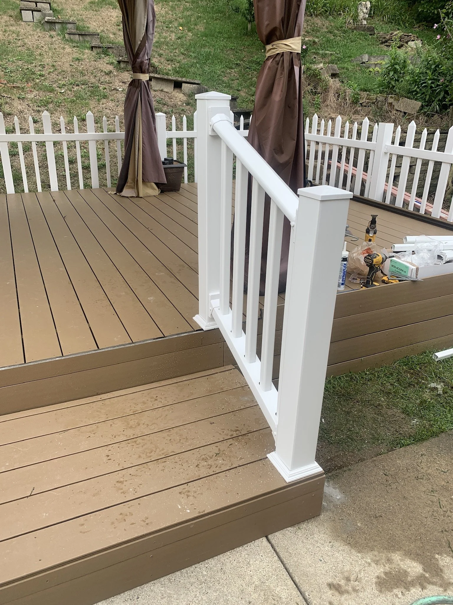 A newly built wooden deck with a white railing and two brown curtains tied back, overlooking a grassy backyard with flower bushes and a stone staircase.