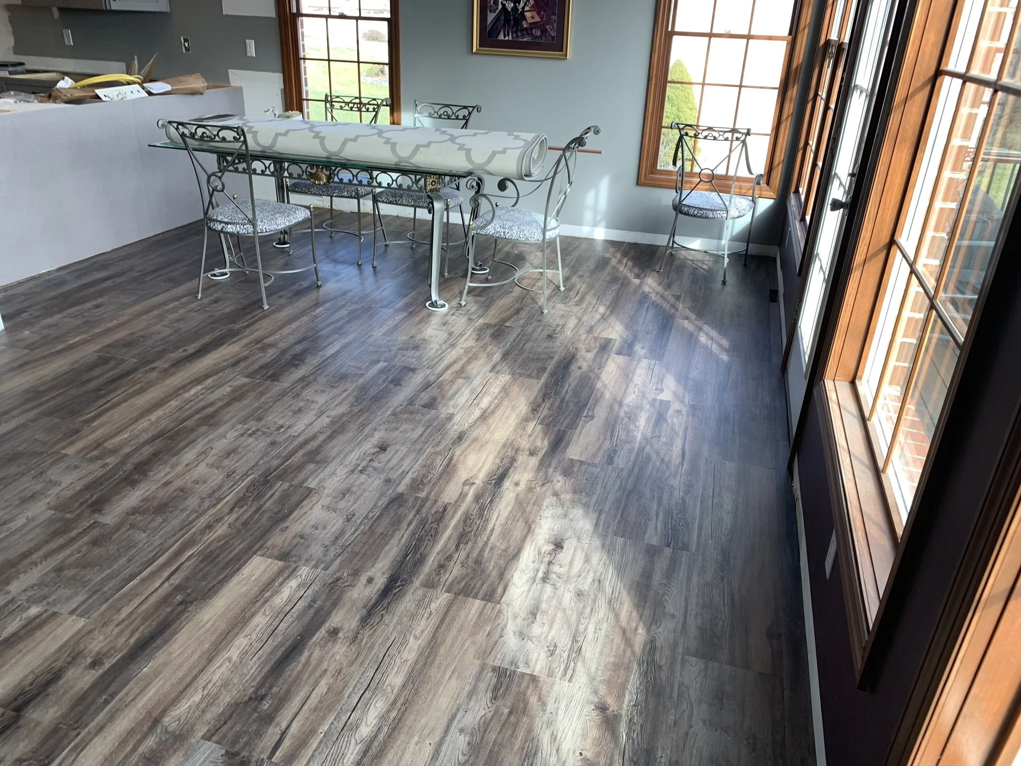 A dining area with a hardwood floor, a table covered with a large roll of paper, surrounded by metal chairs. There are three large windows with wooden trim on one side letting in natural light, with chairs placed beneath the windows. The room appears