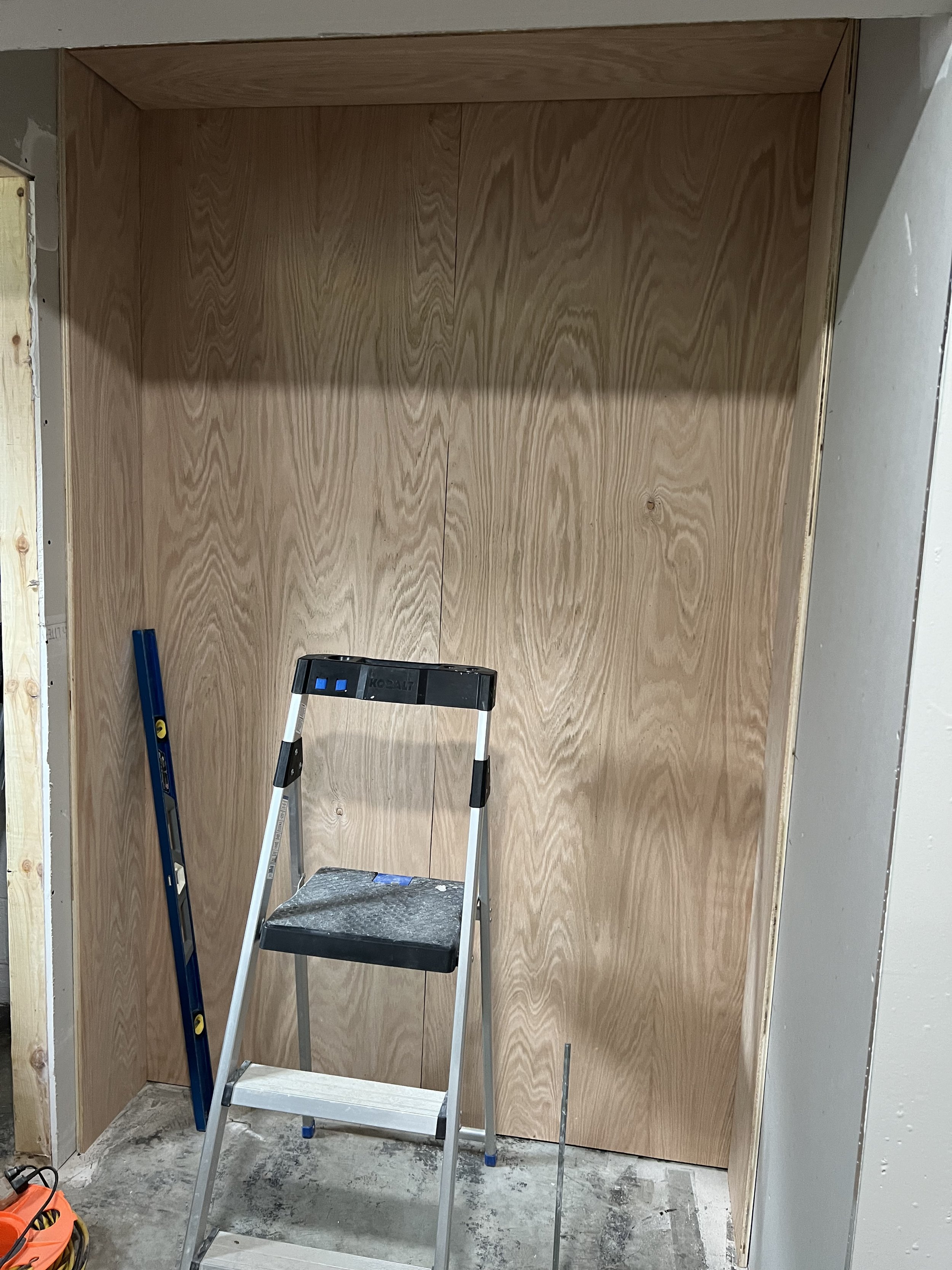 Small wooden corner with a step ladder, a level tool, a measuring tape, and some construction equipment. The wall appears unfinished.
