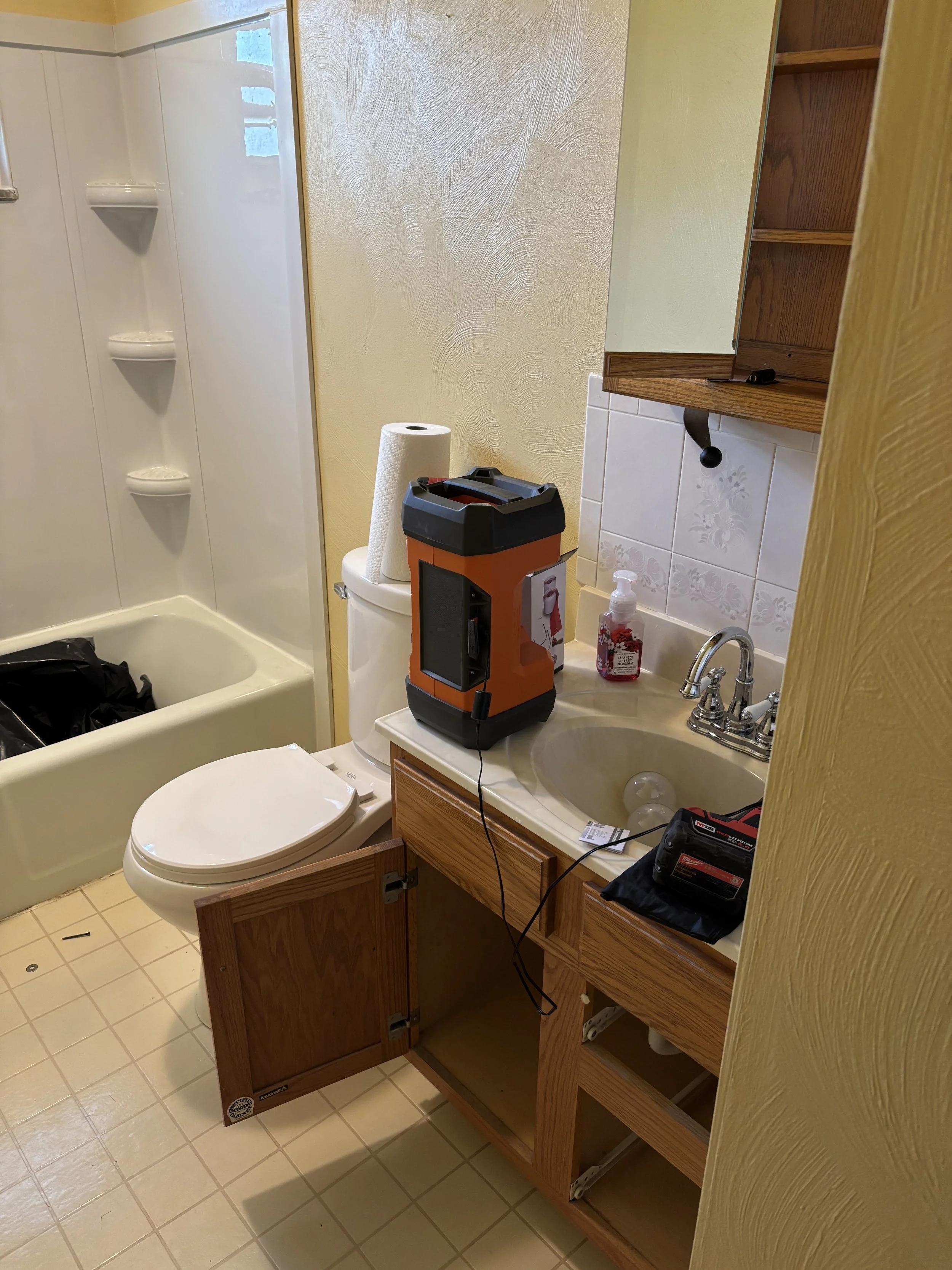 A small bathroom with a vanity, a sink, a toilet, and a bathtub/shower combination. The bathroom countertop has a portable device, a bottle of hand sanitizer, and a small black bag. The bathroom door is open, revealing the cabinet below the sink and 