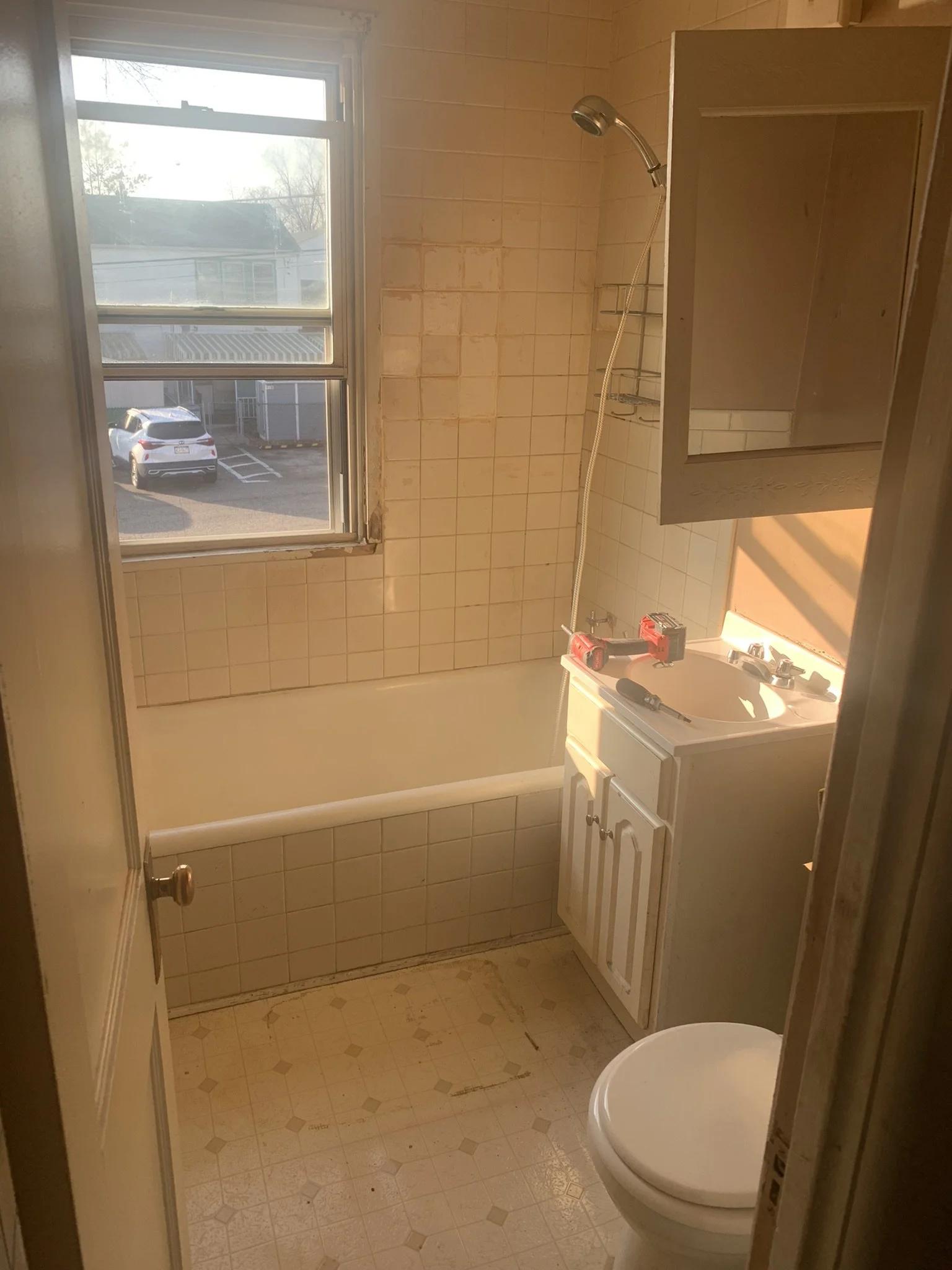 A small bathroom with a window showing a parking lot and neighboring houses. The bathroom has a bathtub, a sink with a cabinet, a mirror cabinet, a toilet, and tile walls. The wall tiles are stained, and the floor tiles have stains and damage. There 