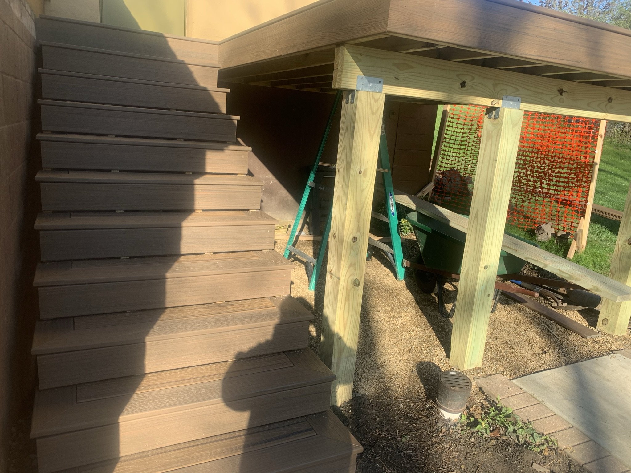 Wooden stairs leading to a deck under construction, with support beams and orange construction fence in the background, and gardening tools and a wheelbarrow underneath.