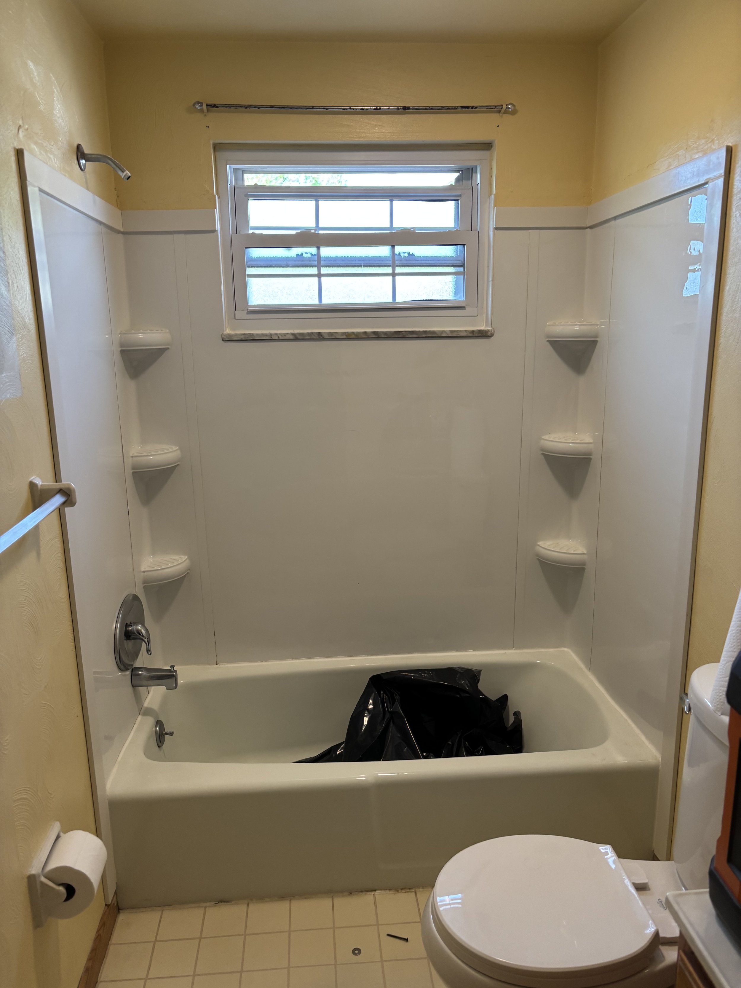Bathroom with a bathtub, wall-mounted shelves, window, toilet, and a black plastic bag inside the bathtub.