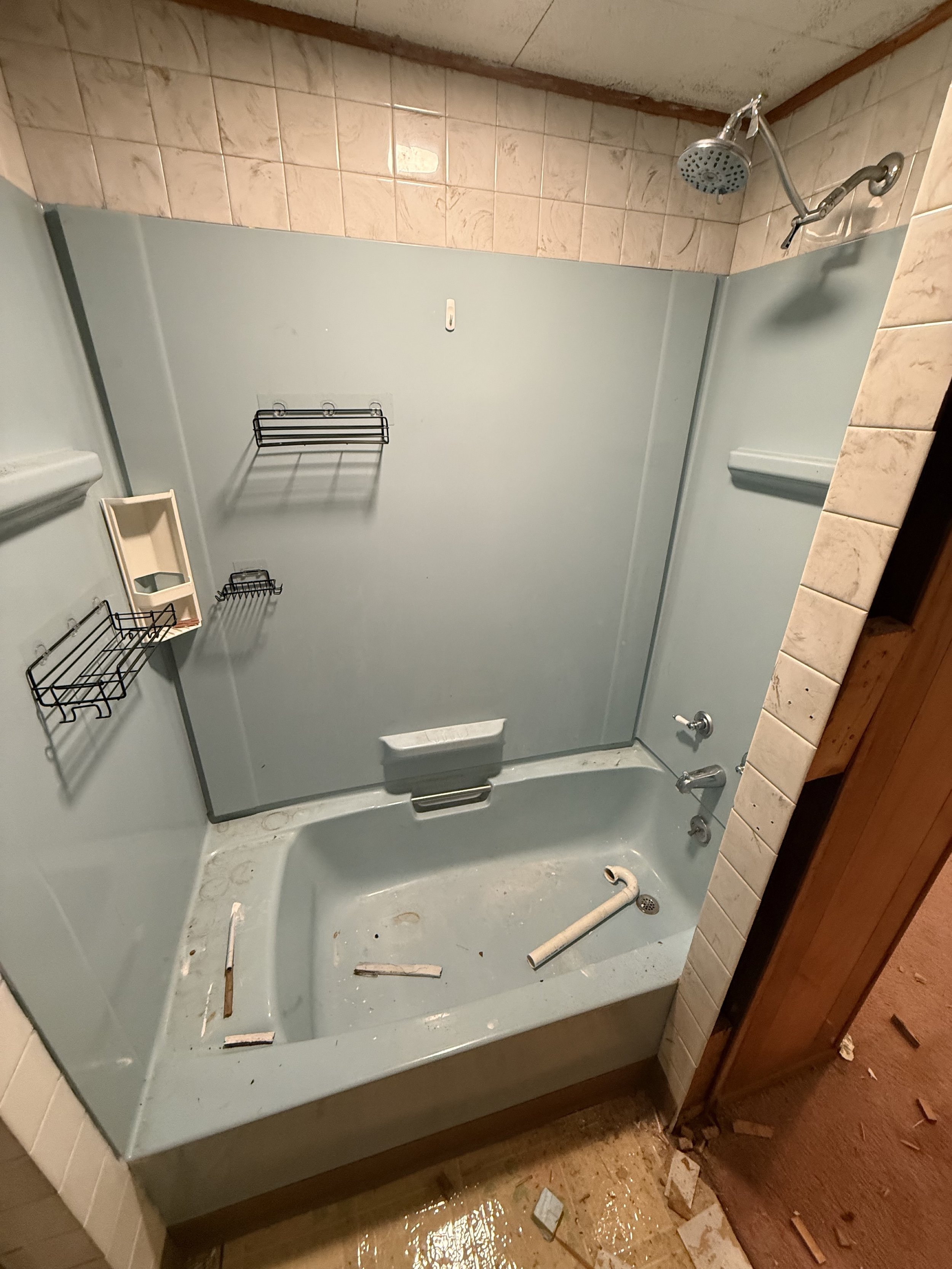 Empty bathtub with debris, rust, and disconnected plumbing in a bathroom shower area. Wall-mounted showerhead, soap dish, and wire racks on the walls.