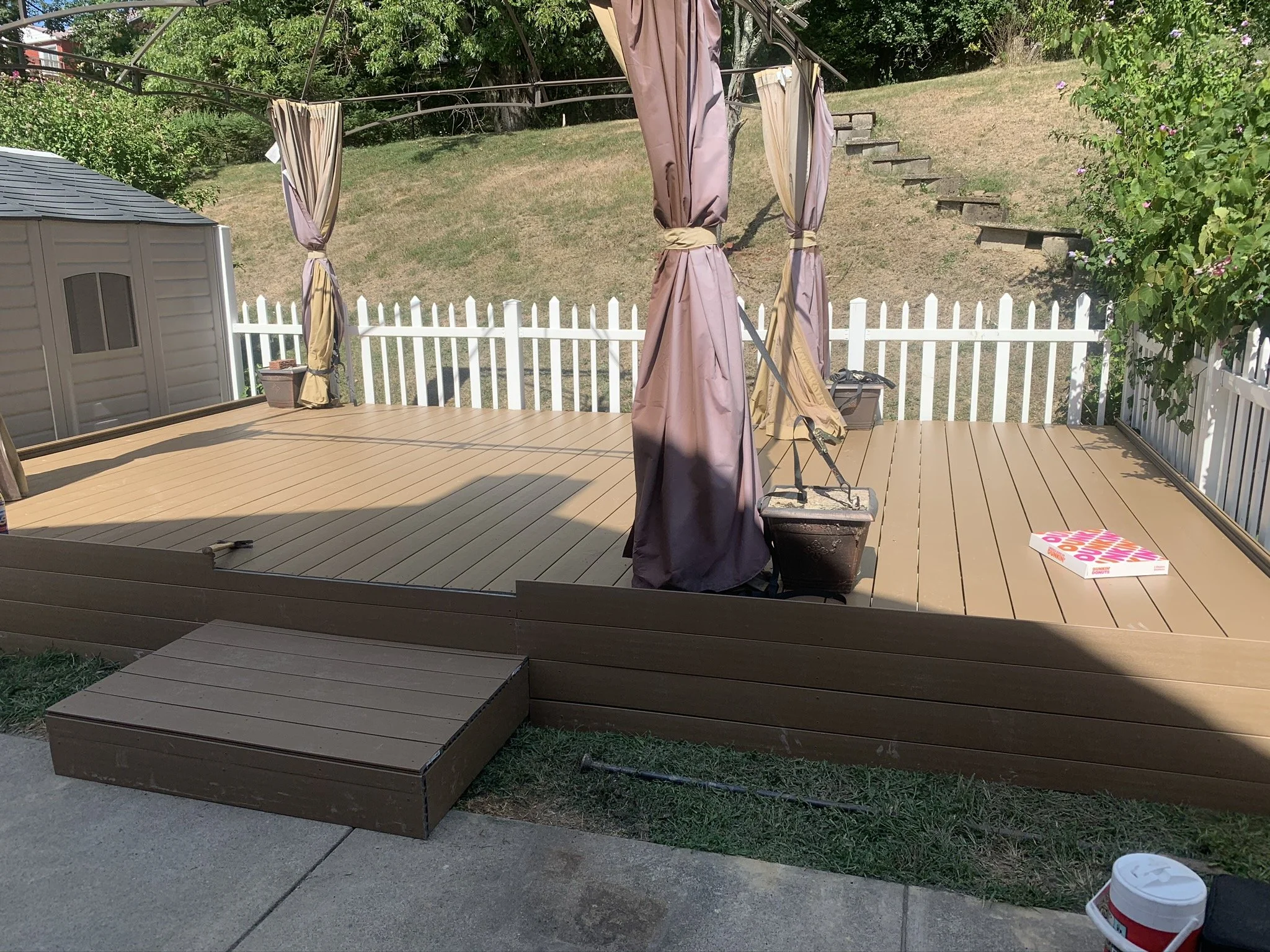 A backyard deck with tan-colored wood flooring, surrounded by a white picket fence. There are four purple and tan umbrellas (two closed, two partially open) on the deck, with a small patio table and some items like a book or magazine and a container.