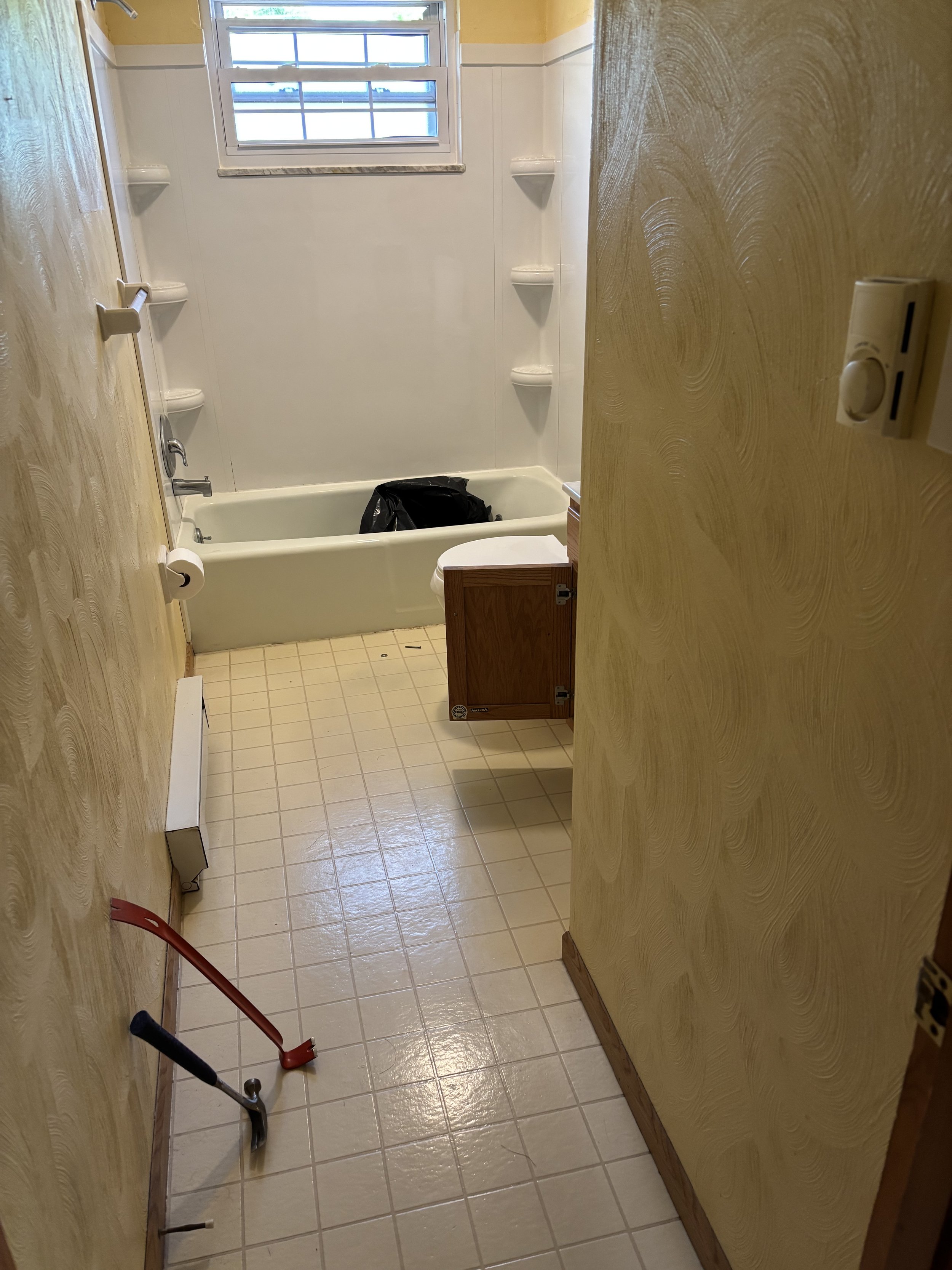 A small bathroom with yellow textured walls and beige tiled floor. There is a bathtub with a black trash bag inside, a window above the tub, and a small wooden cabinet attached to the wall. A red crowbar and a wrench are on the floor near the entry.